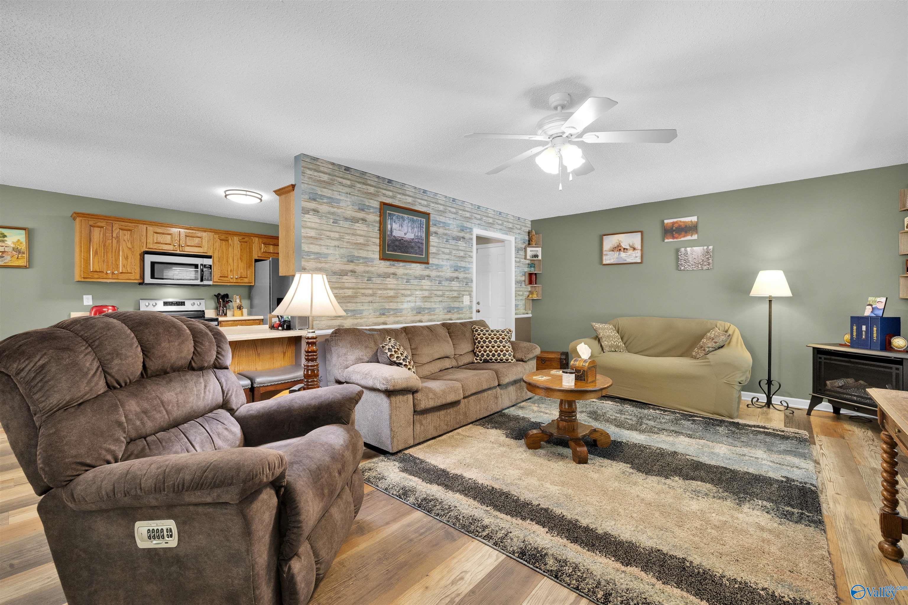 Furnished living room with a rustic plank accent wall, reclining sofas, and an open kitchen.