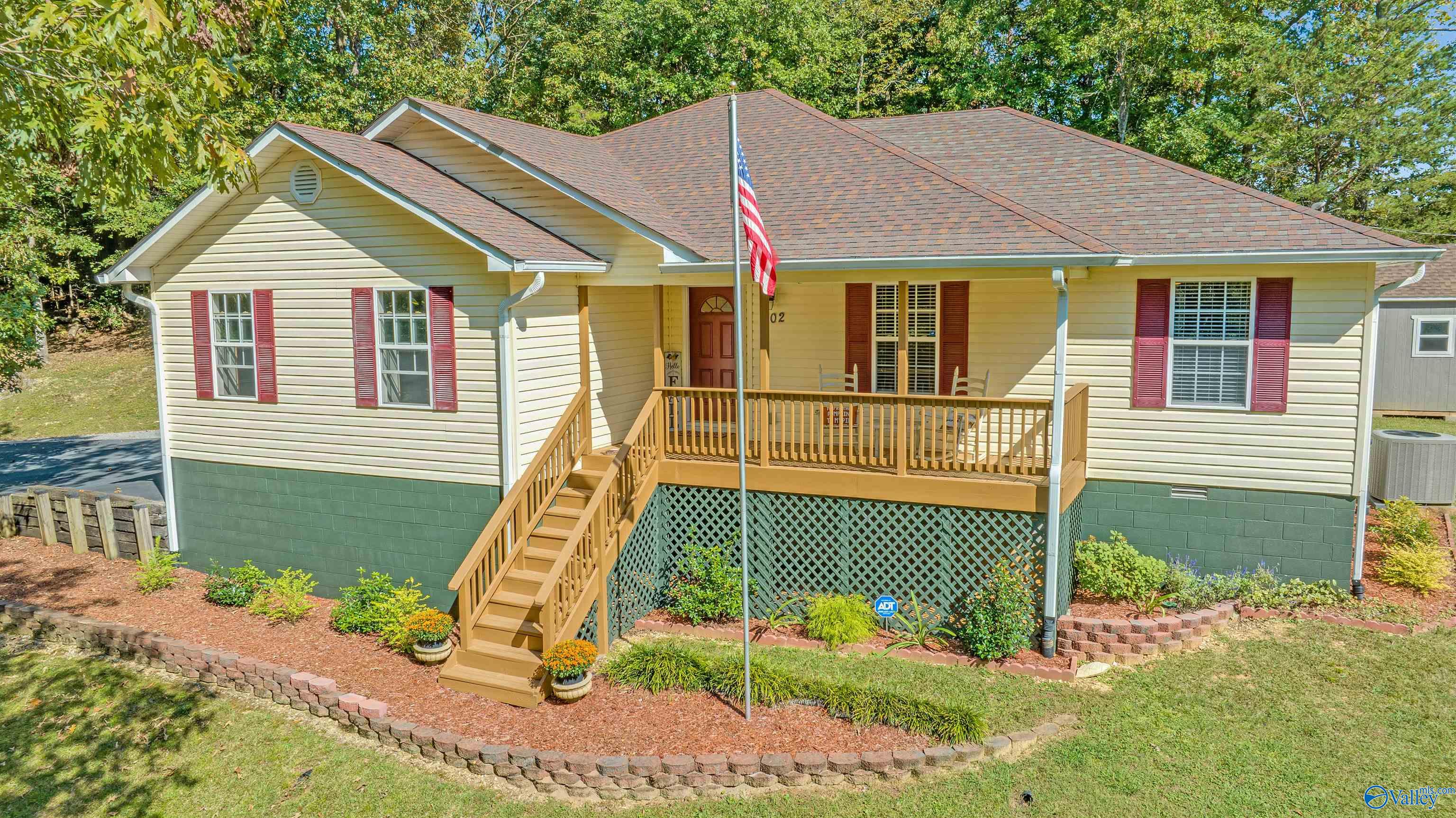 Cream-sided home with burgundy shutters, raised wooden porch, flower beds, and an American flag.