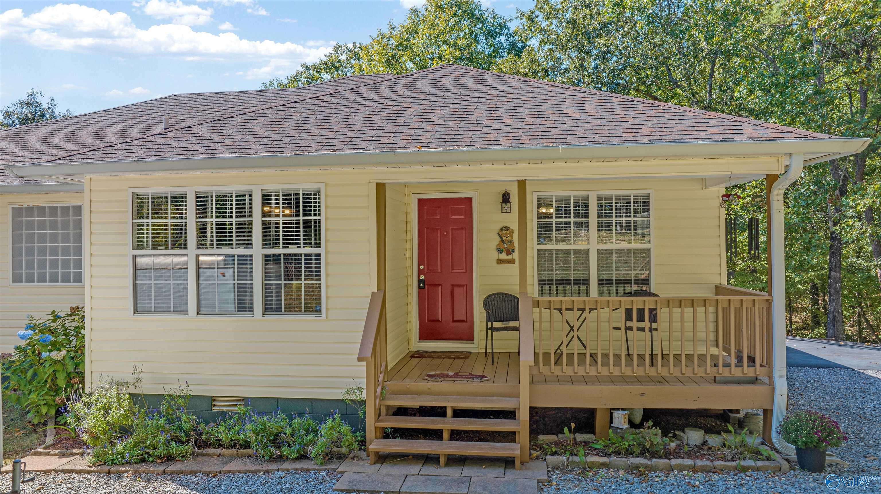 Pale yellow house with a covered wooden porch, red front door, and mature trees nearby.
