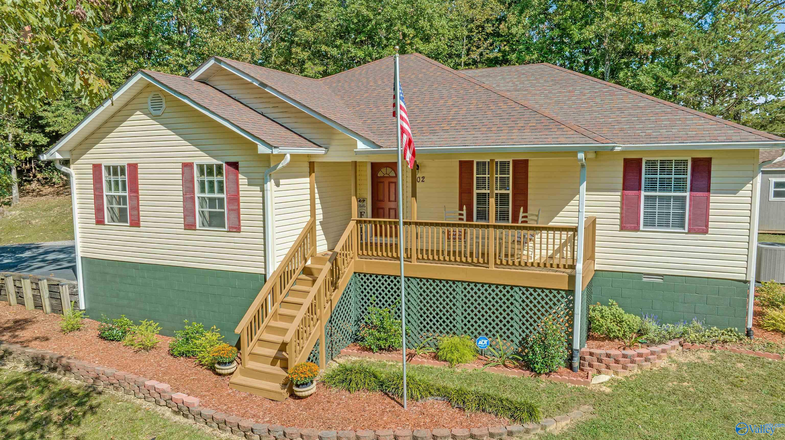 Beige house with red shutters, raised wooden porch and stairs, and landscaped garden beds.