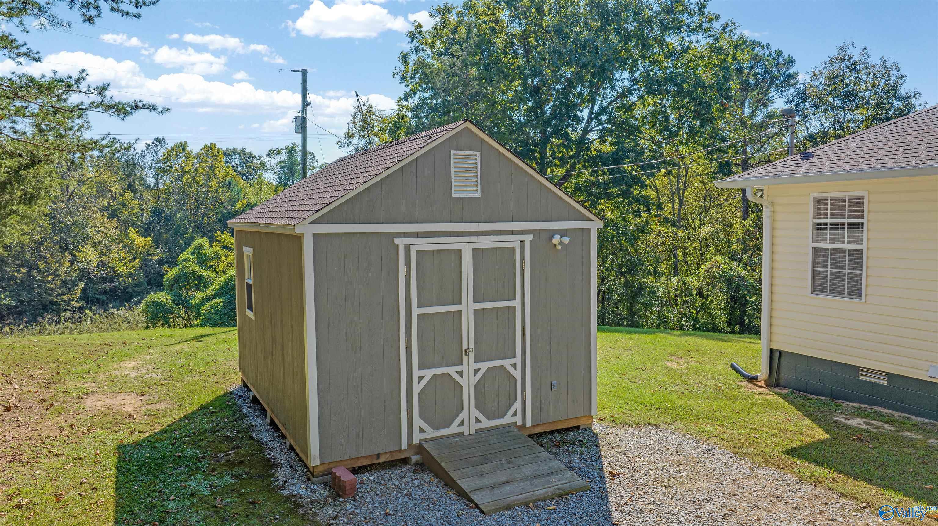 Gray storage shed with white-trimmed double doors and a wooden ramp beside a house.