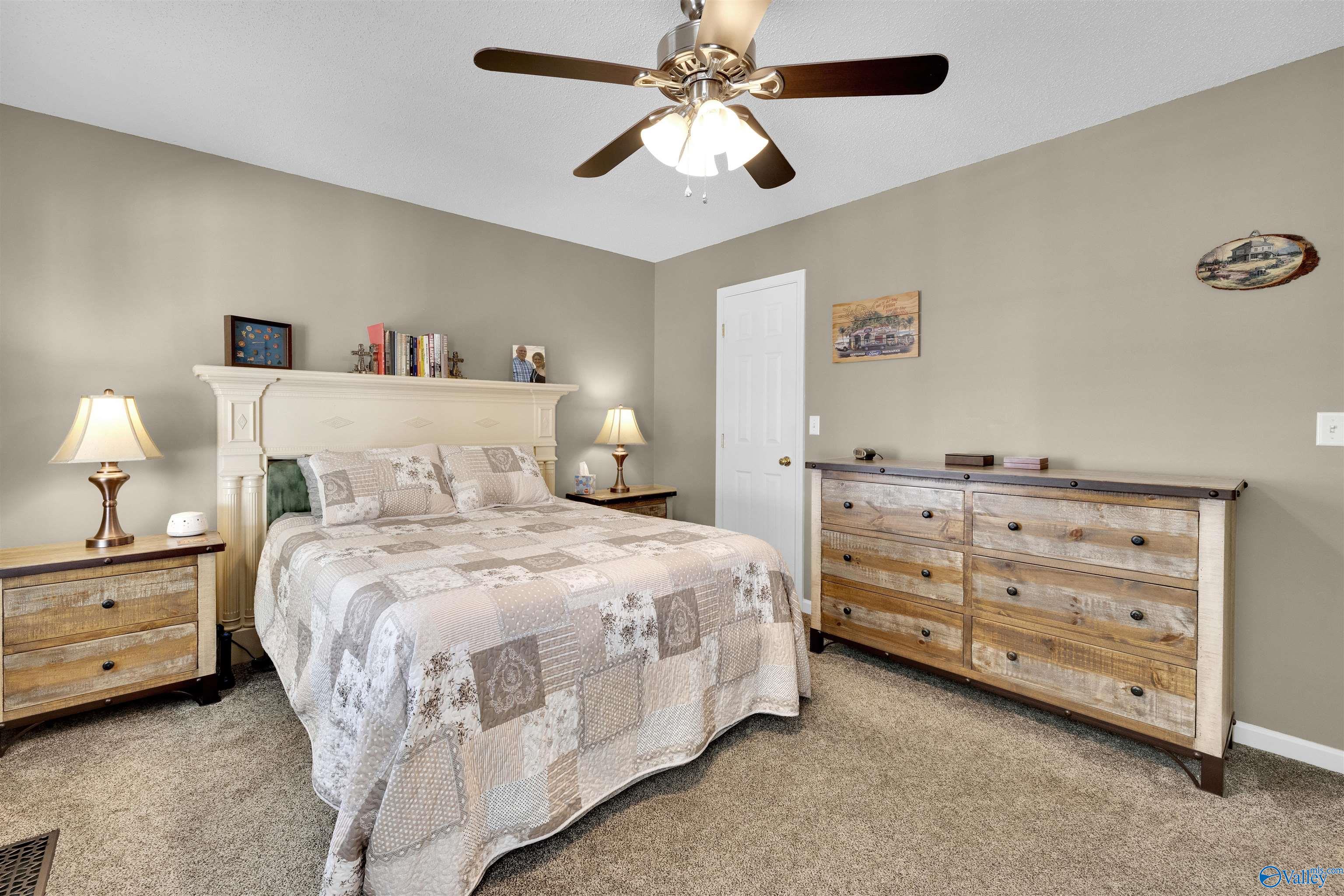Furnished bedroom with a patchwork quilt, carved cream headboard, rustic wood dresser, and ceiling fan.