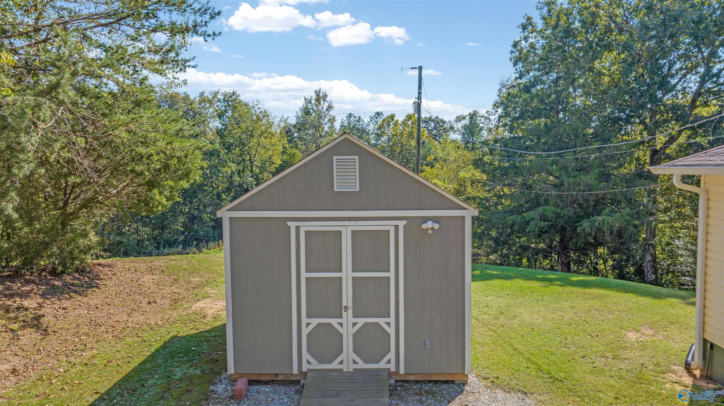 Gray storage shed with white trim and double doors, set on a lawn among mature trees.