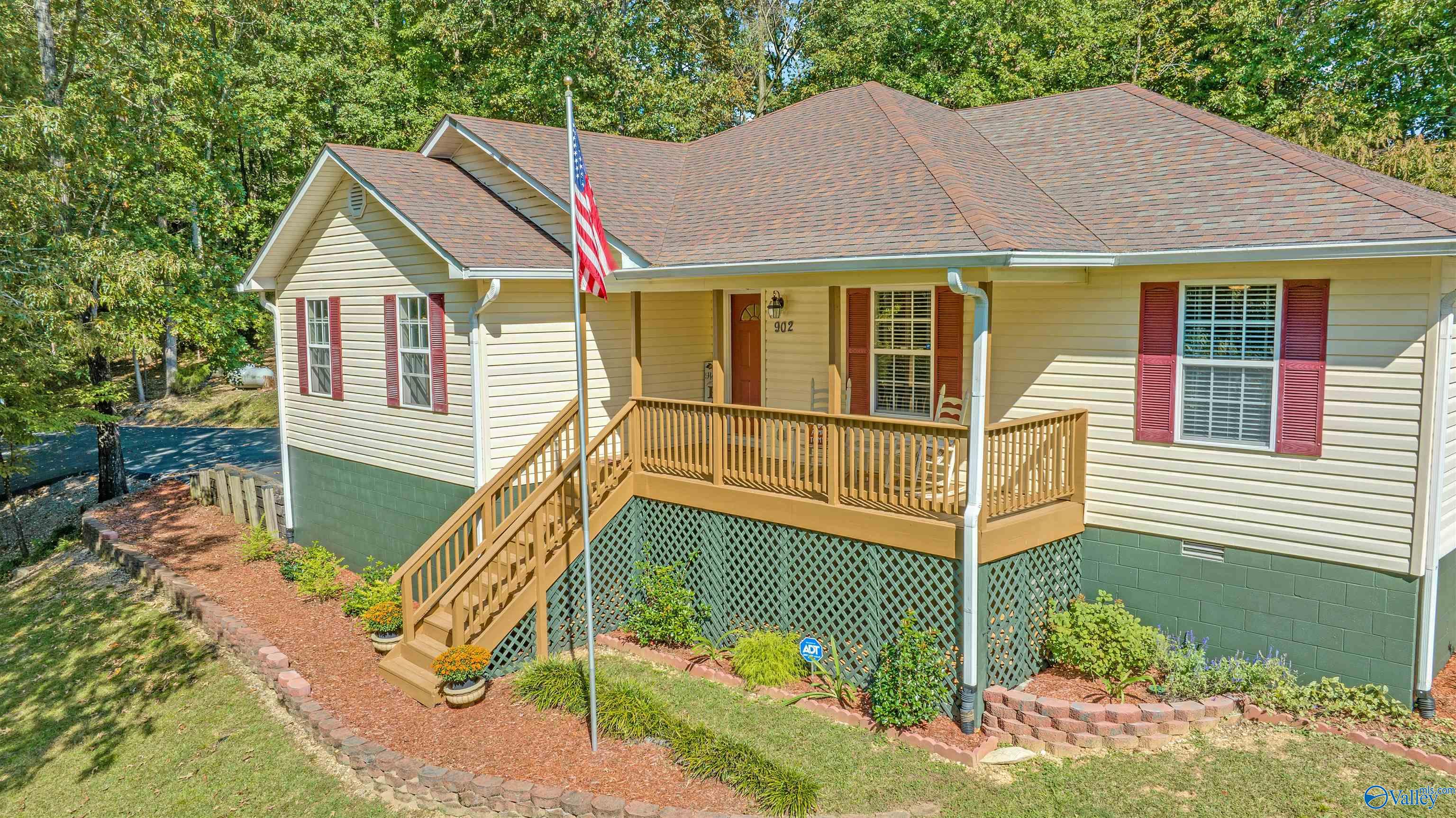 Home exterior with a raised wooden porch, red shutters, landscaped beds, and an American flag.