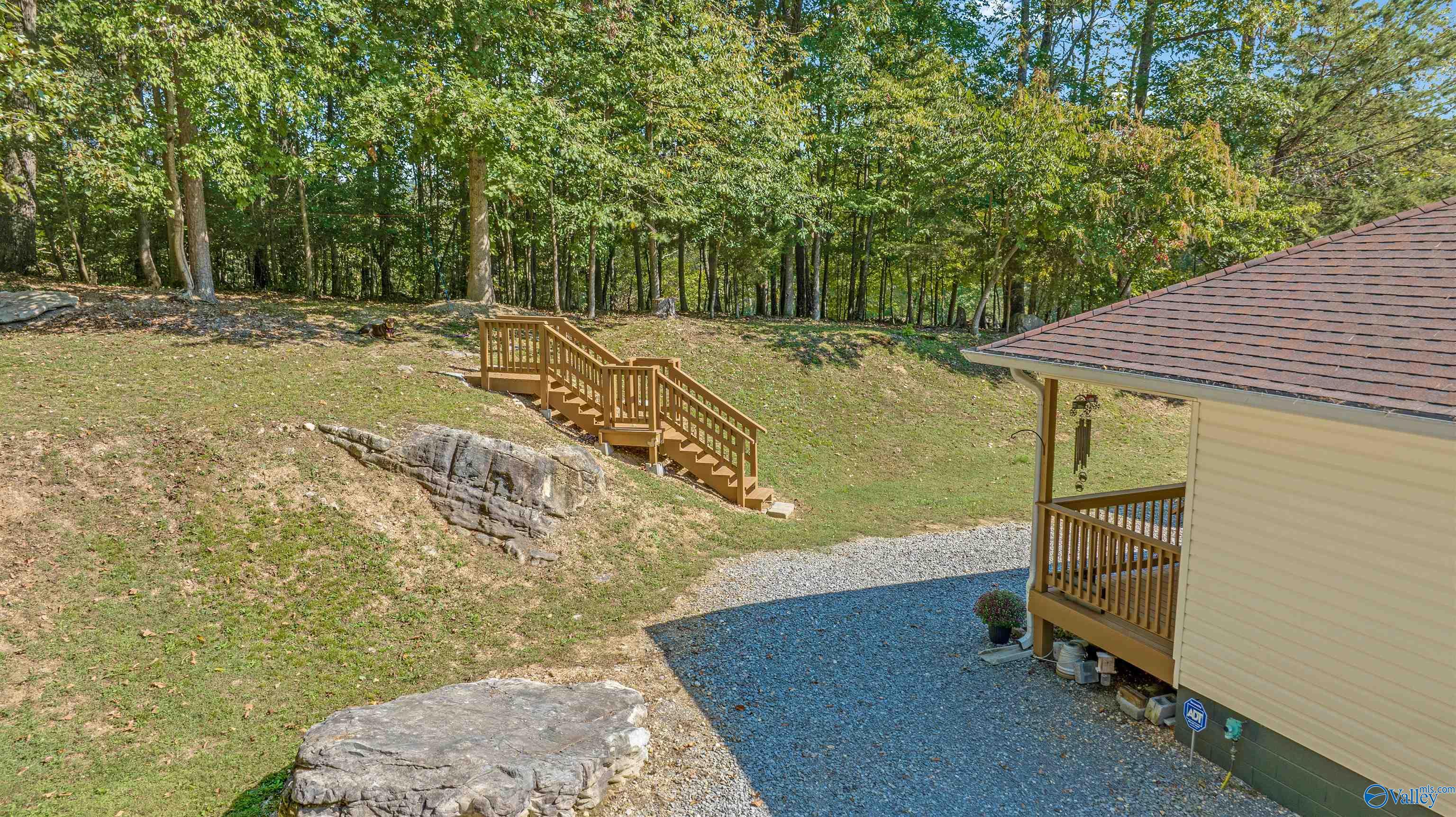 Wooded sloping yard with a wooden staircase, exposed rock ledges, and gravel beside the house.