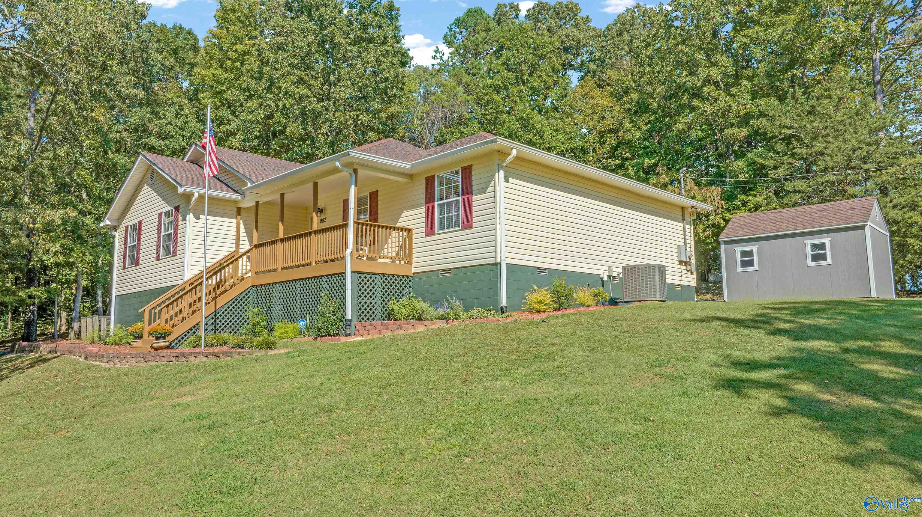House with a raised wooden porch and American flag, beside a detached gray shed on a sloping lawn.
