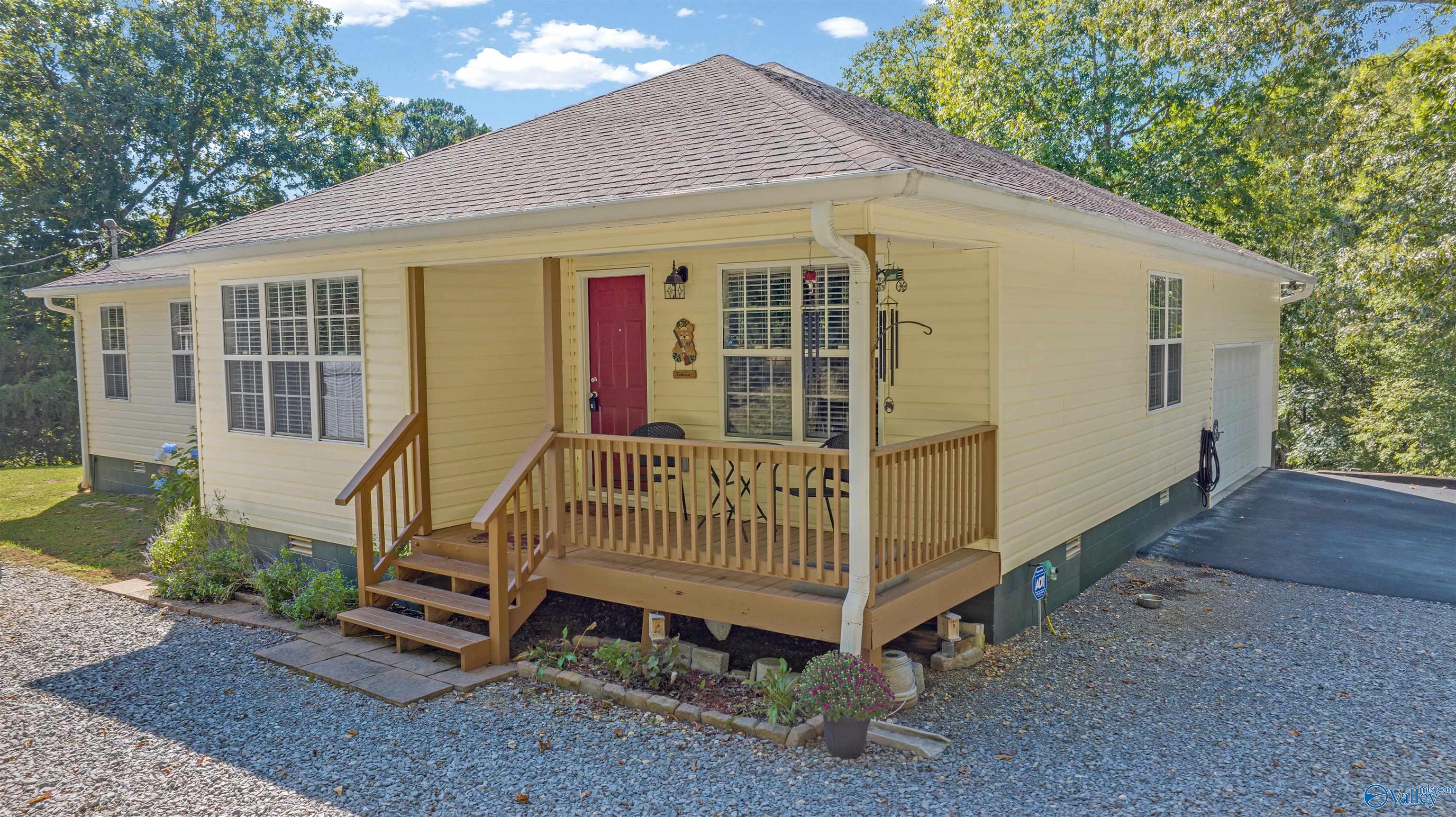 Cream-colored home with a red front door and covered wooden porch surrounded by gravel and trees.