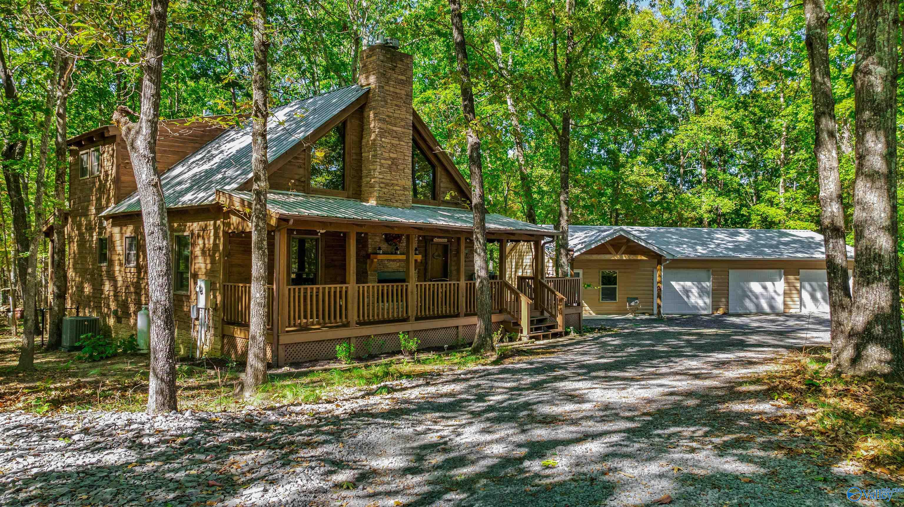 Wood-sided cabin with a stone chimney, covered porch, and detached garage in a leafy woodland setting.