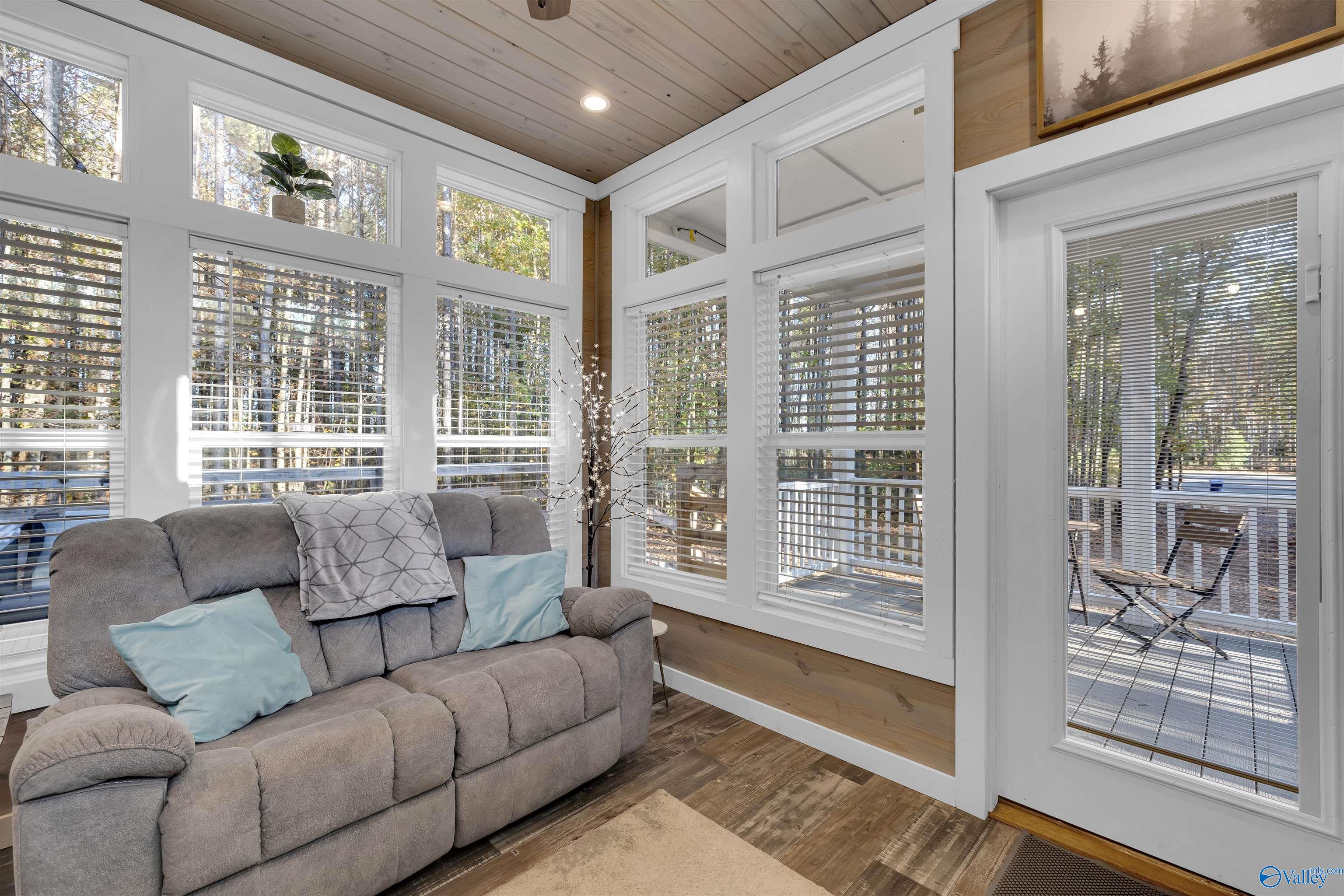 Living room with a gray reclining sofa, tall windows, wood ceiling, and wooded views.