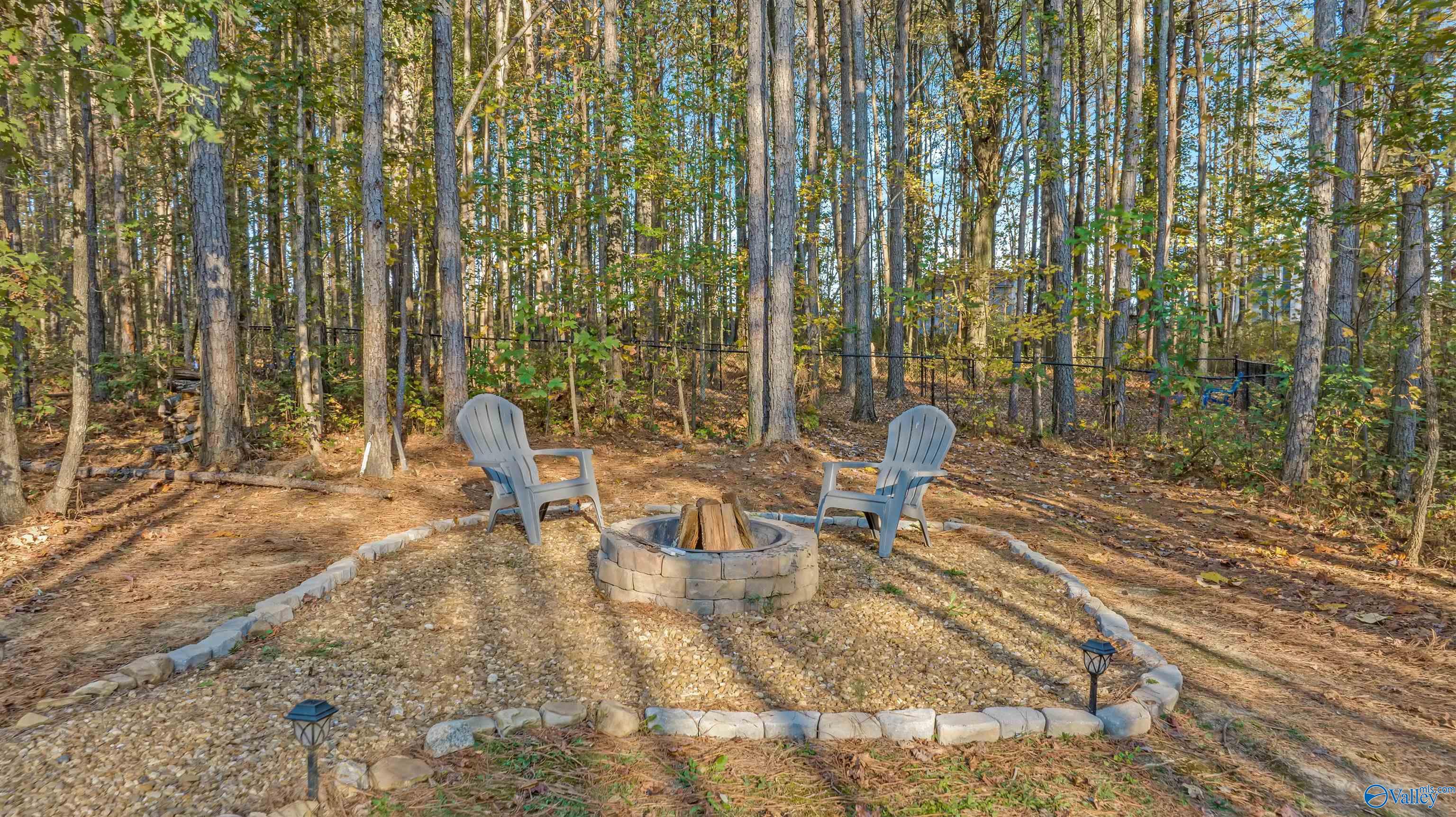 Wooded lot with a stone fire pit and two Adirondack chairs in a gravel clearing.