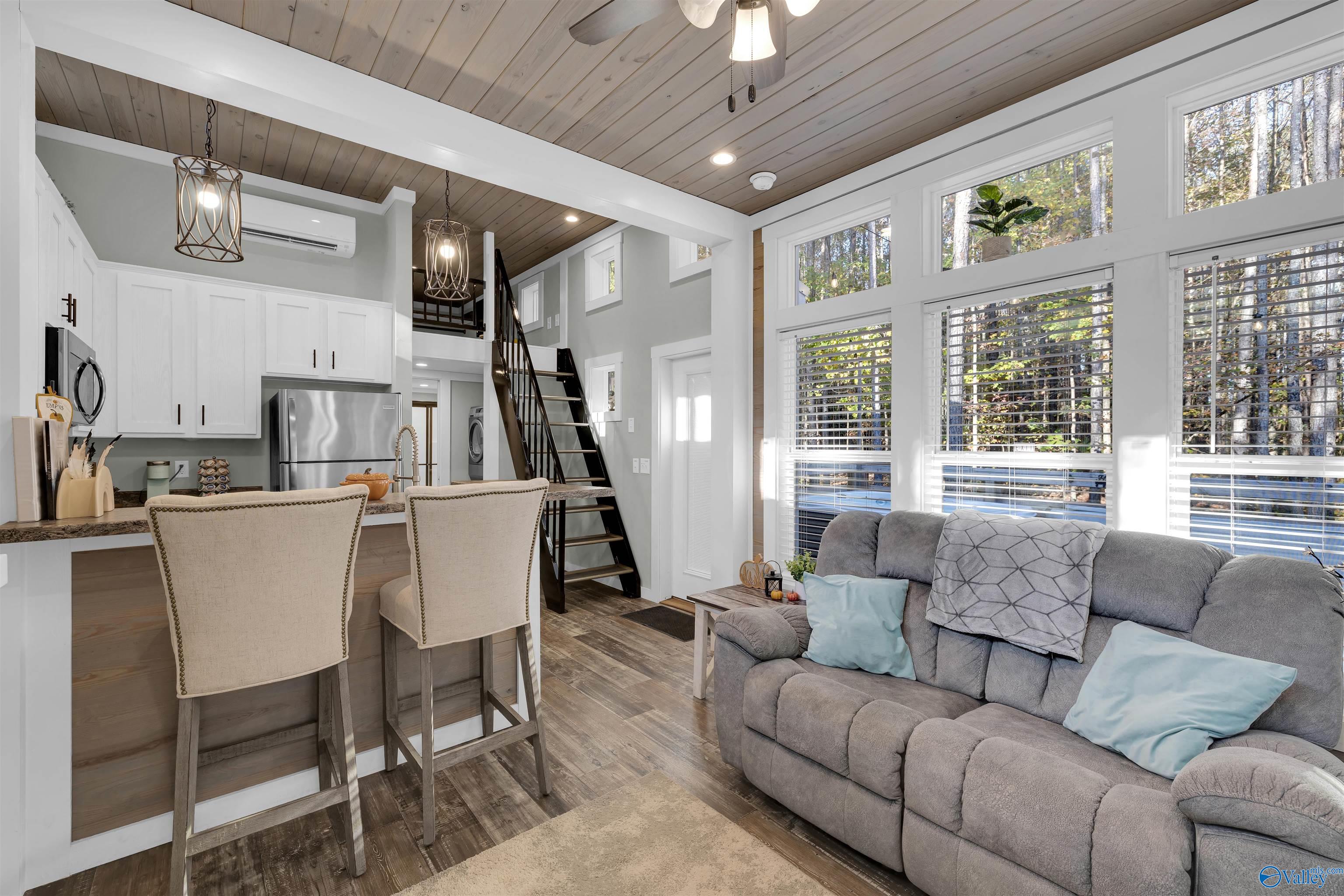 Open-plan living room and kitchen with a gray sectional, breakfast bar, and tall wooded-view windows.