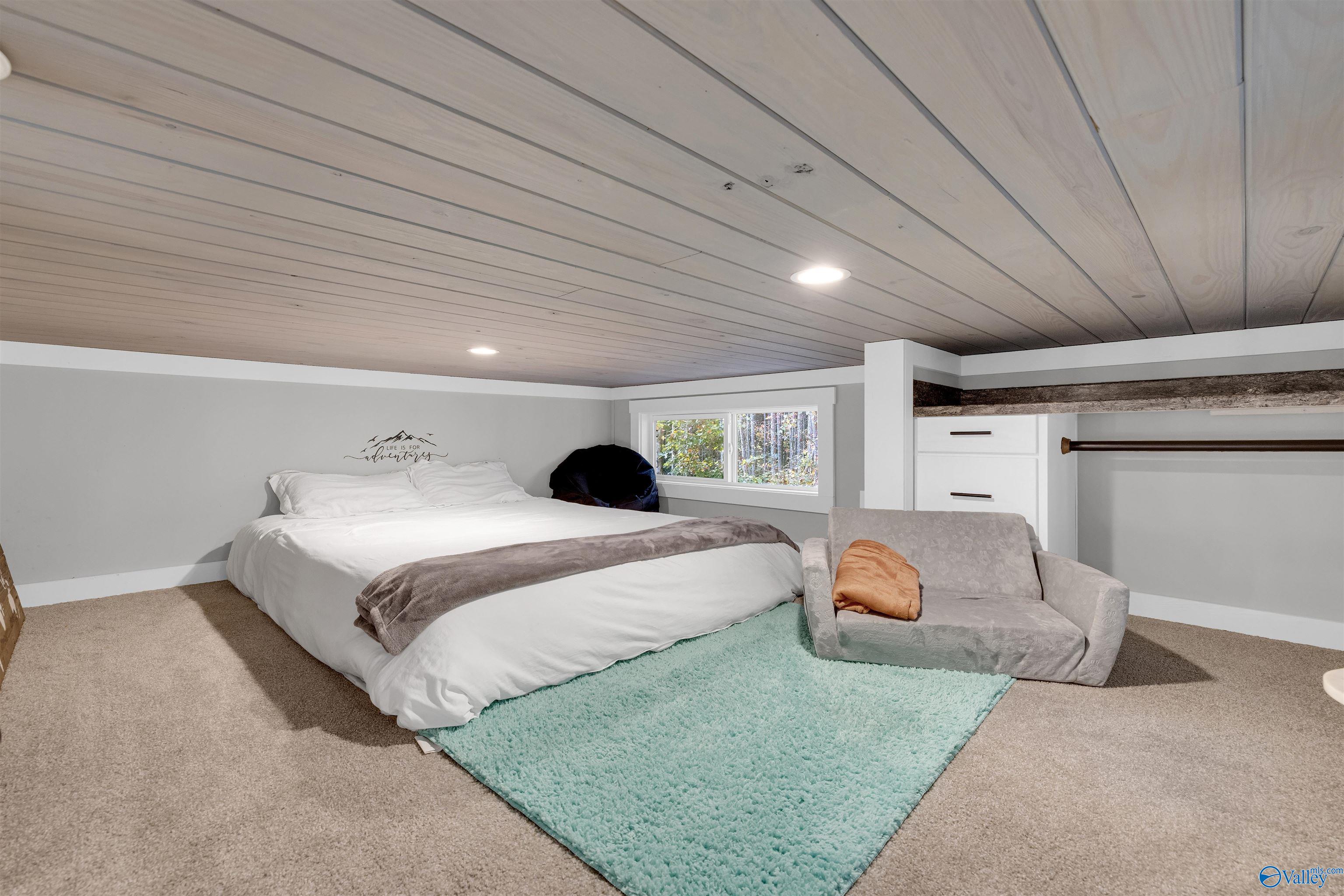 Furnished bedroom with a white bed, turquoise rug, and light wood plank ceiling.