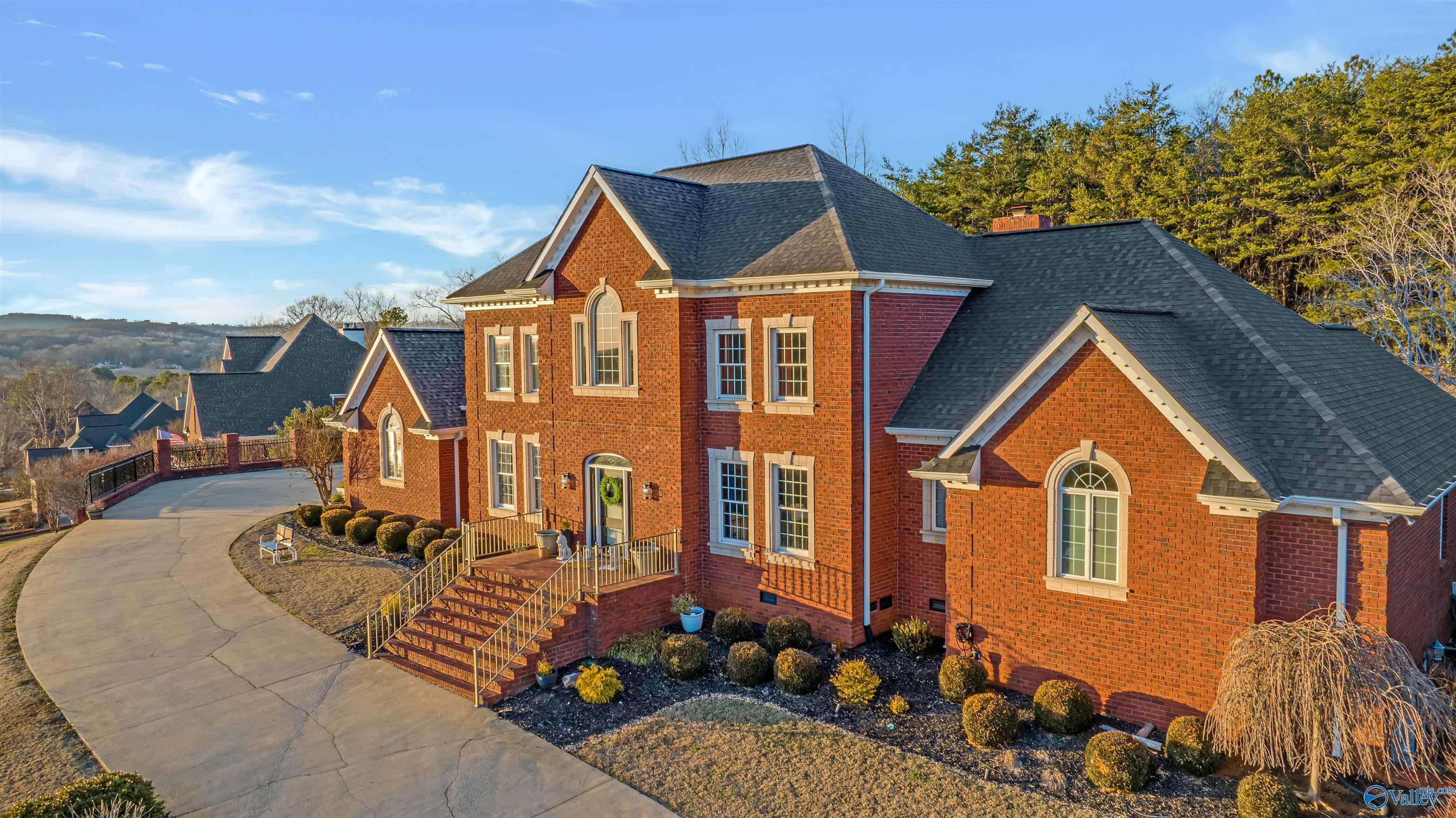Two-story red brick home with multiple gables, landscaped shrubs, and a curving driveway.