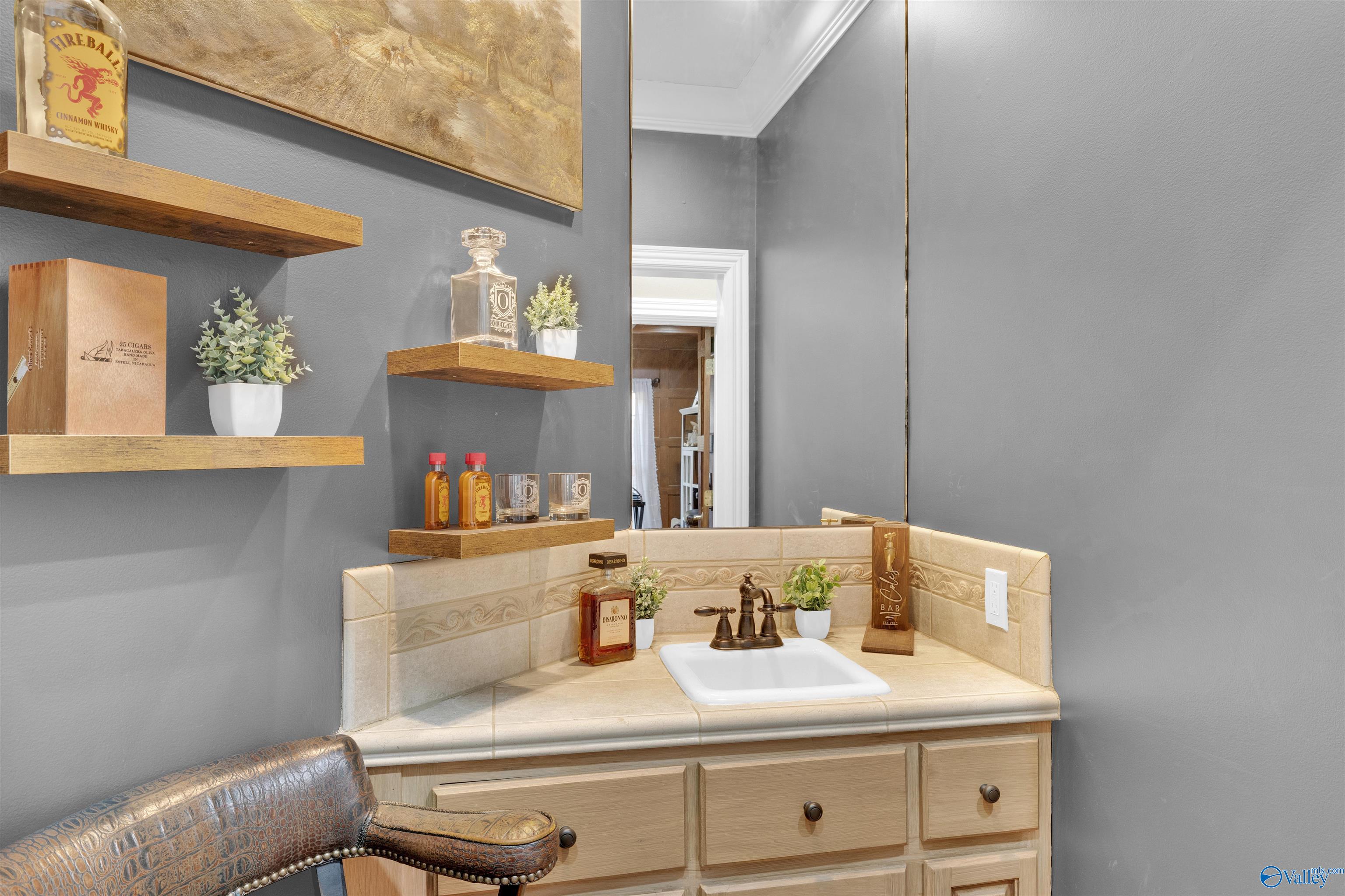 Bathroom vanity with beige tile, a large mirror, floating display shelves, and a patterned leather chair.