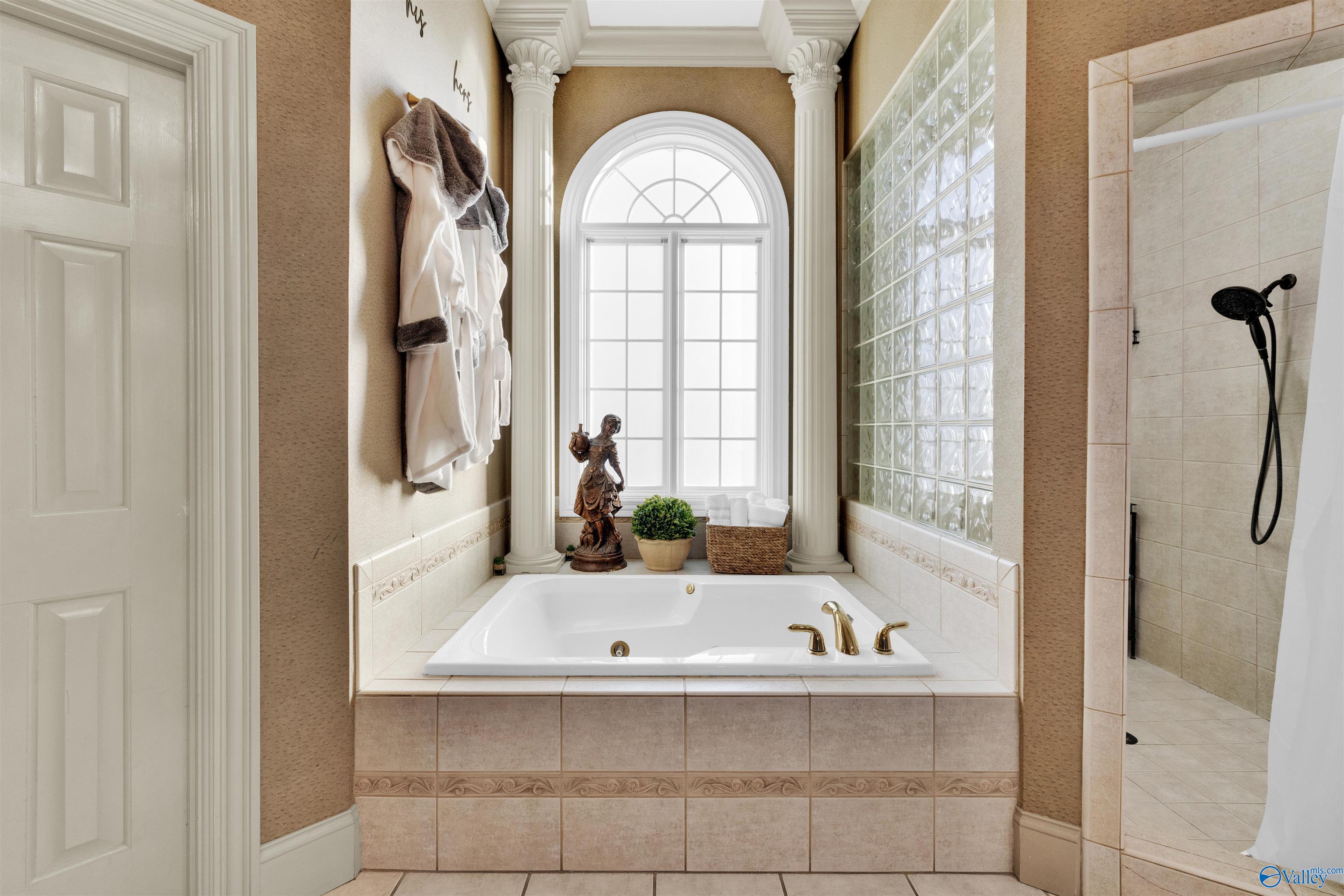 Bathroom with a built-in soaking tub beneath an arched window, beside a glass-block wall and tiled shower.