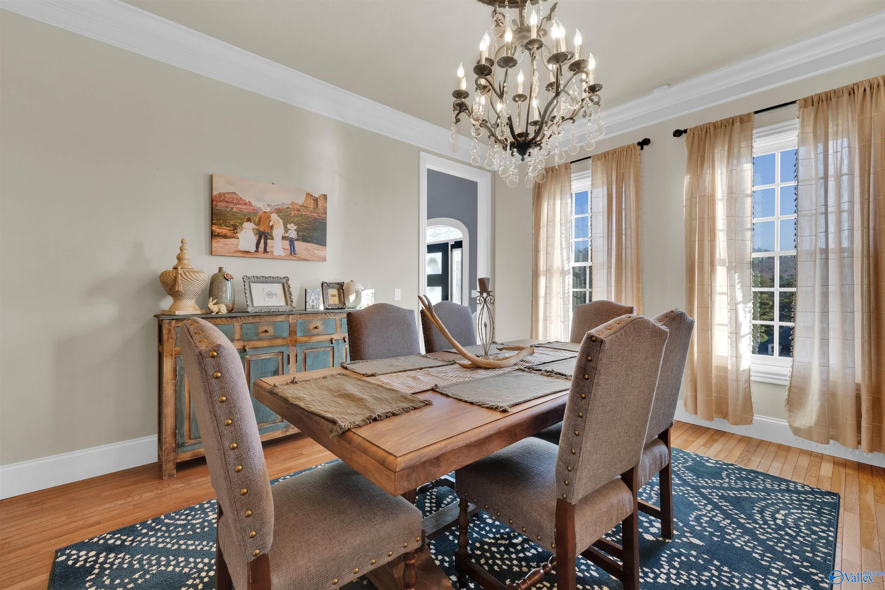 Dining room with a crystal chandelier, wooden table, upholstered chairs, and tall curtained windows.