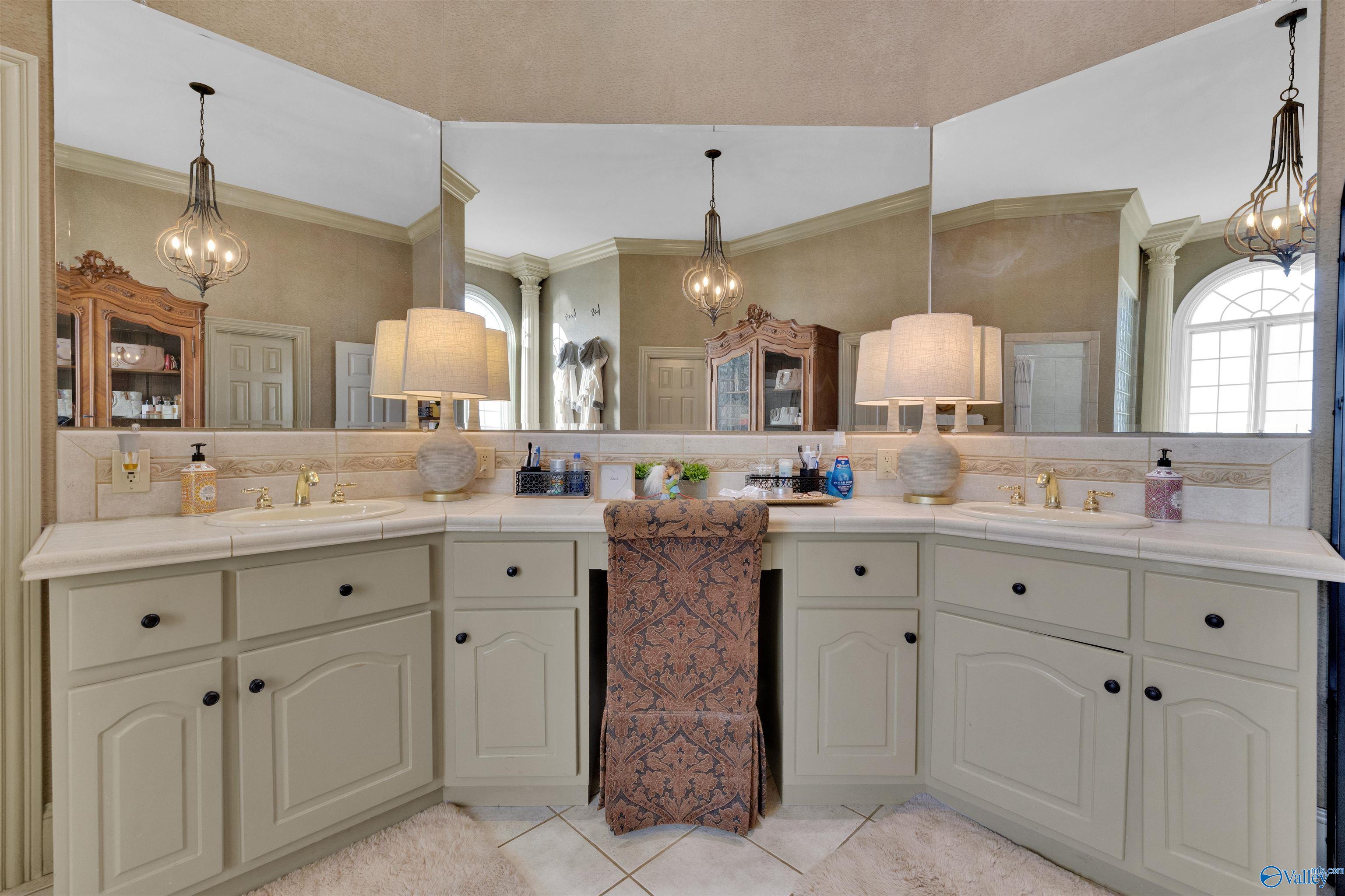 Bathroom with a cream double-sink vanity, wall-to-wall mirrors, and a patterned chair.