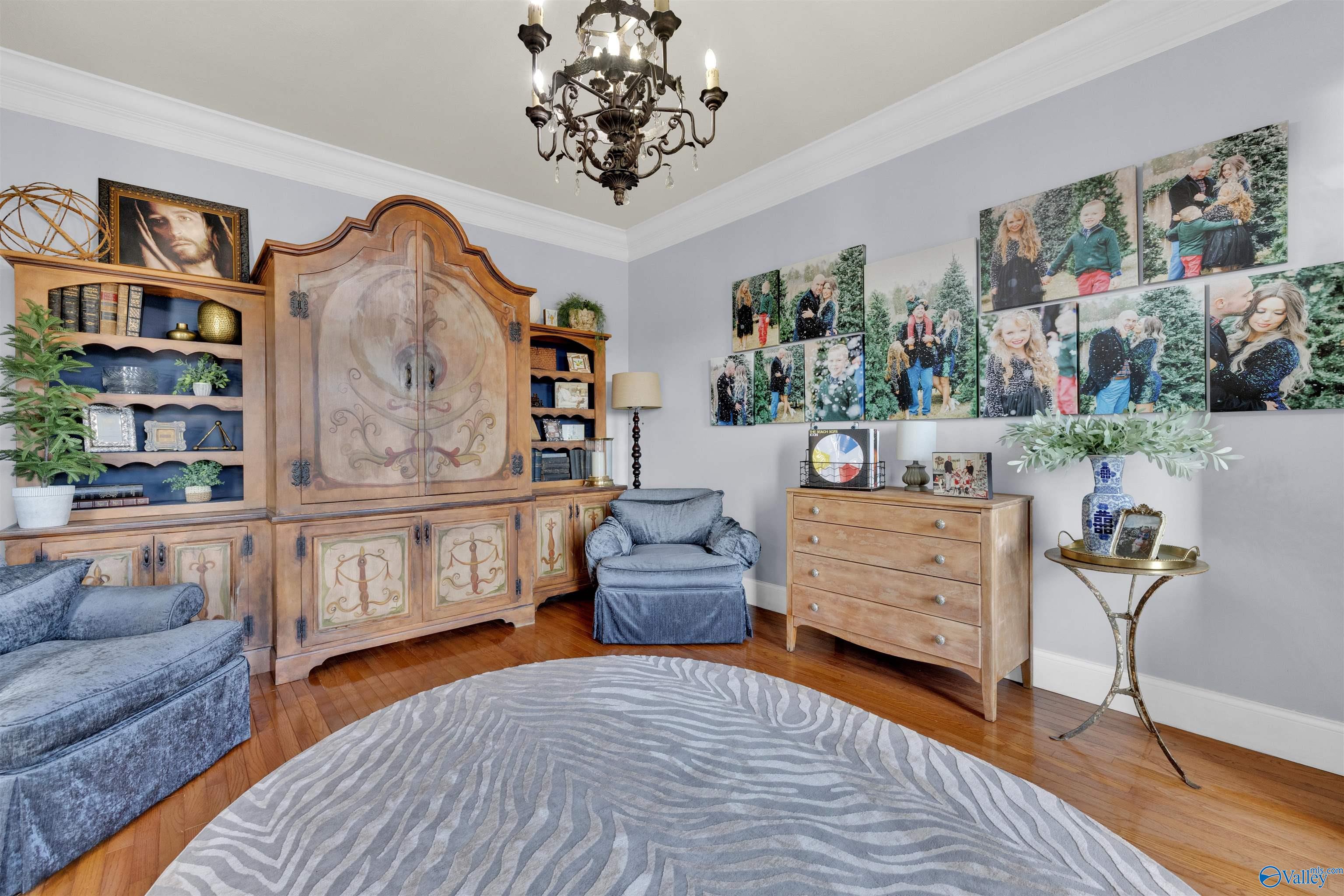 Furnished sitting room with an ornate wooden cabinet, blue chairs, hardwood floors, and a gallery of family portraits.
