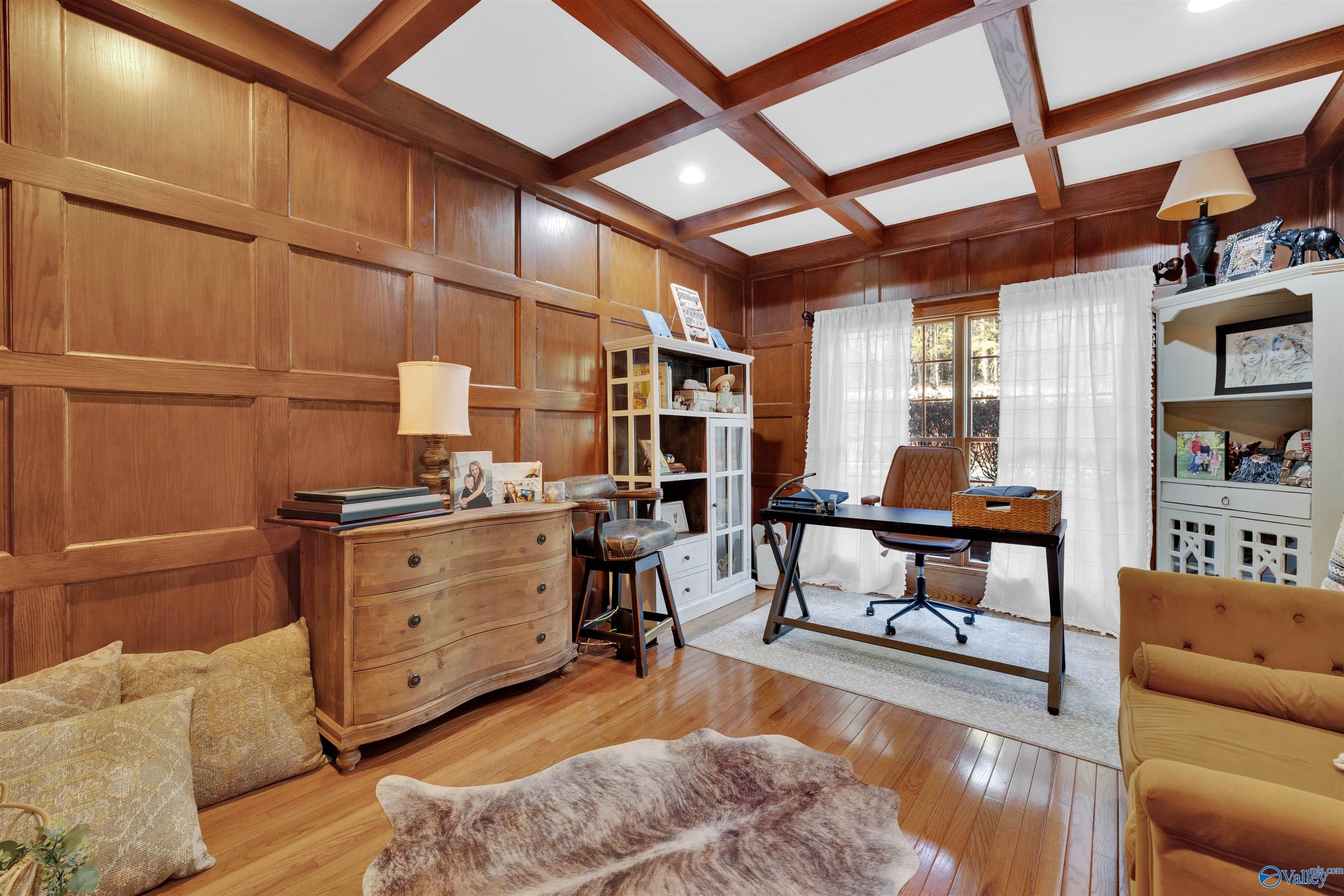 Wood-paneled home office with coffered ceiling, desk, built-in shelving, and a curtained window.