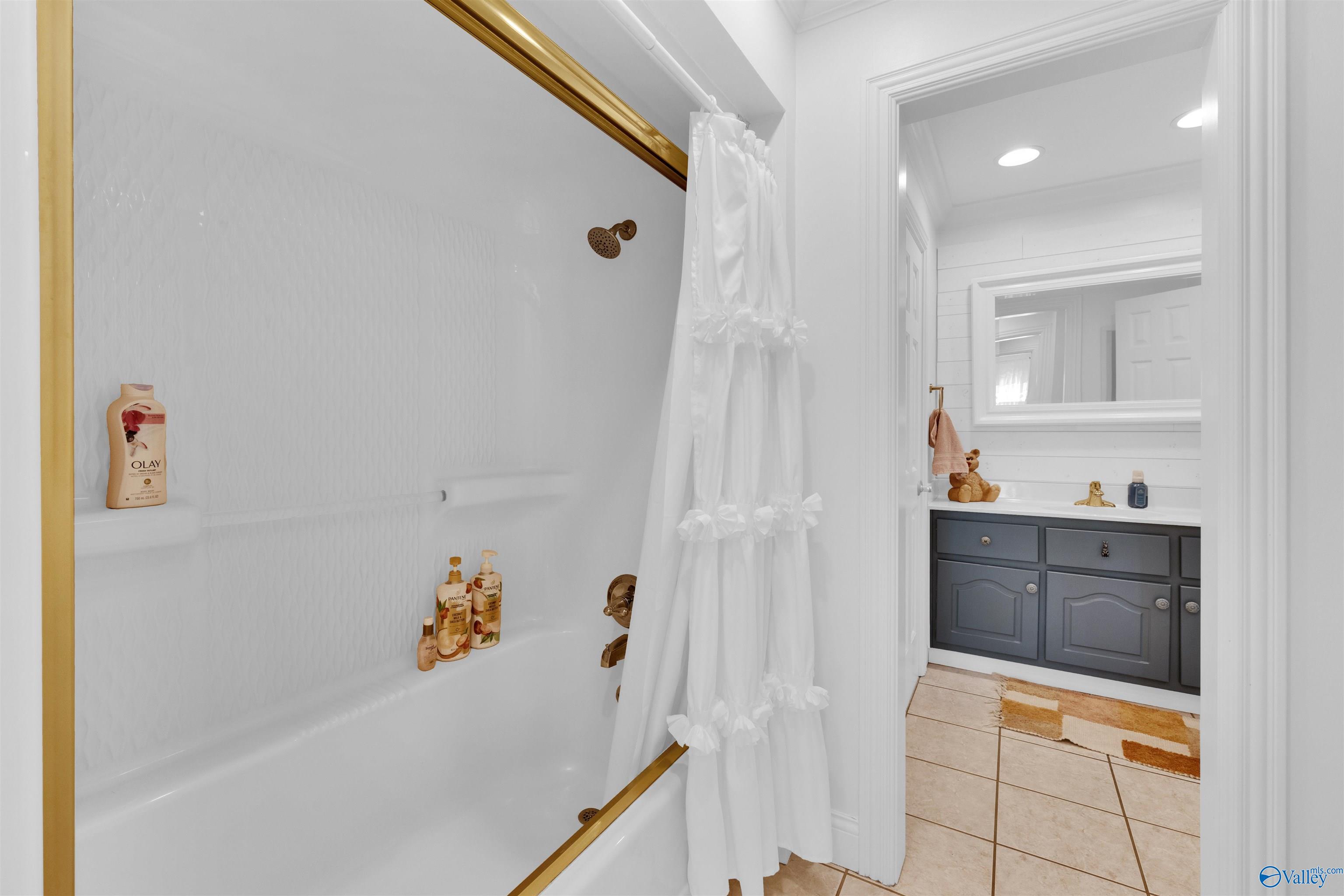 Bathroom with a gold-trimmed bathtub and white ruffled curtain, looking through to a gray vanity.