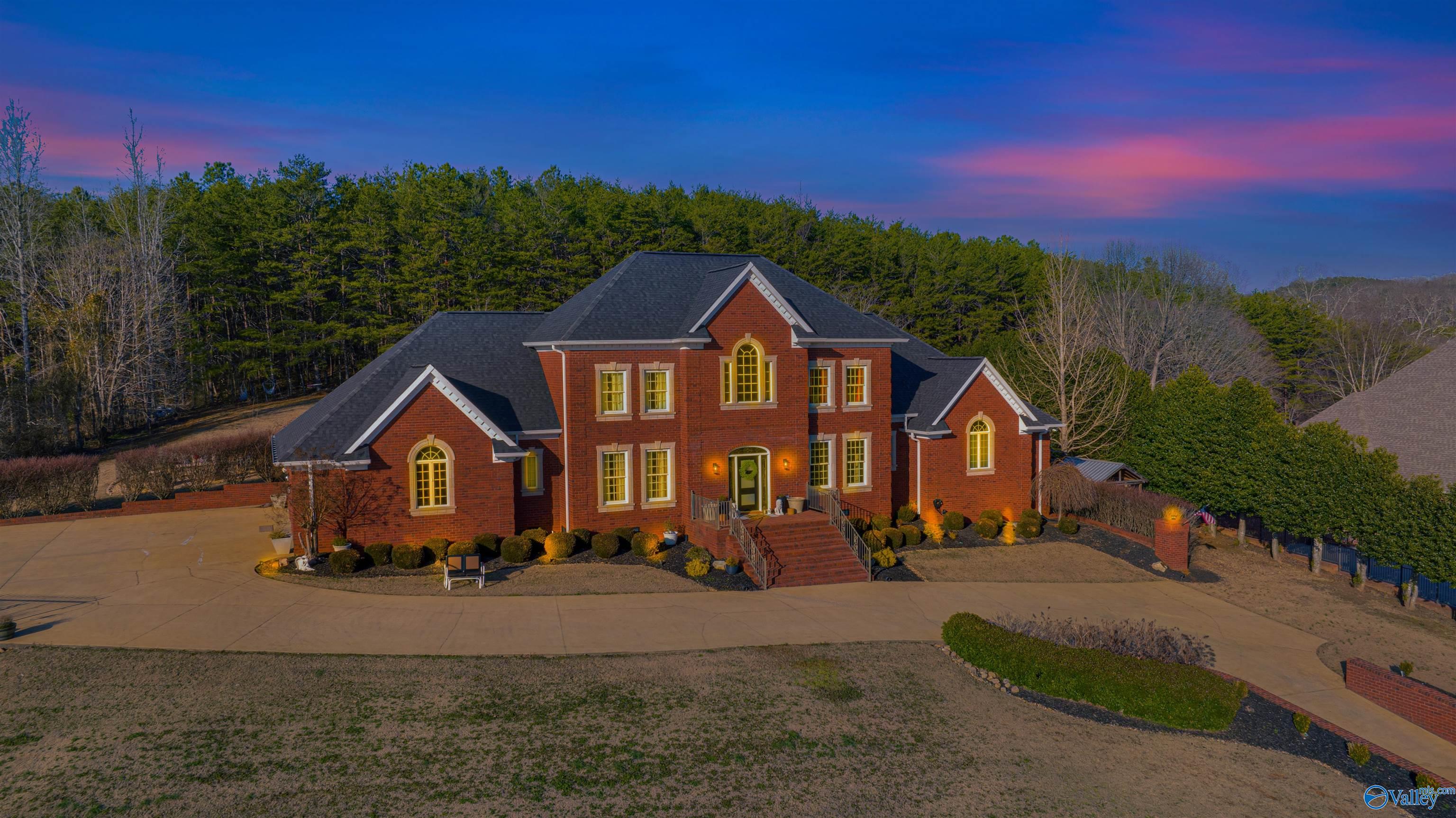 Two-story red-brick house with illuminated windows, landscaped grounds, and a curved driveway at dusk.