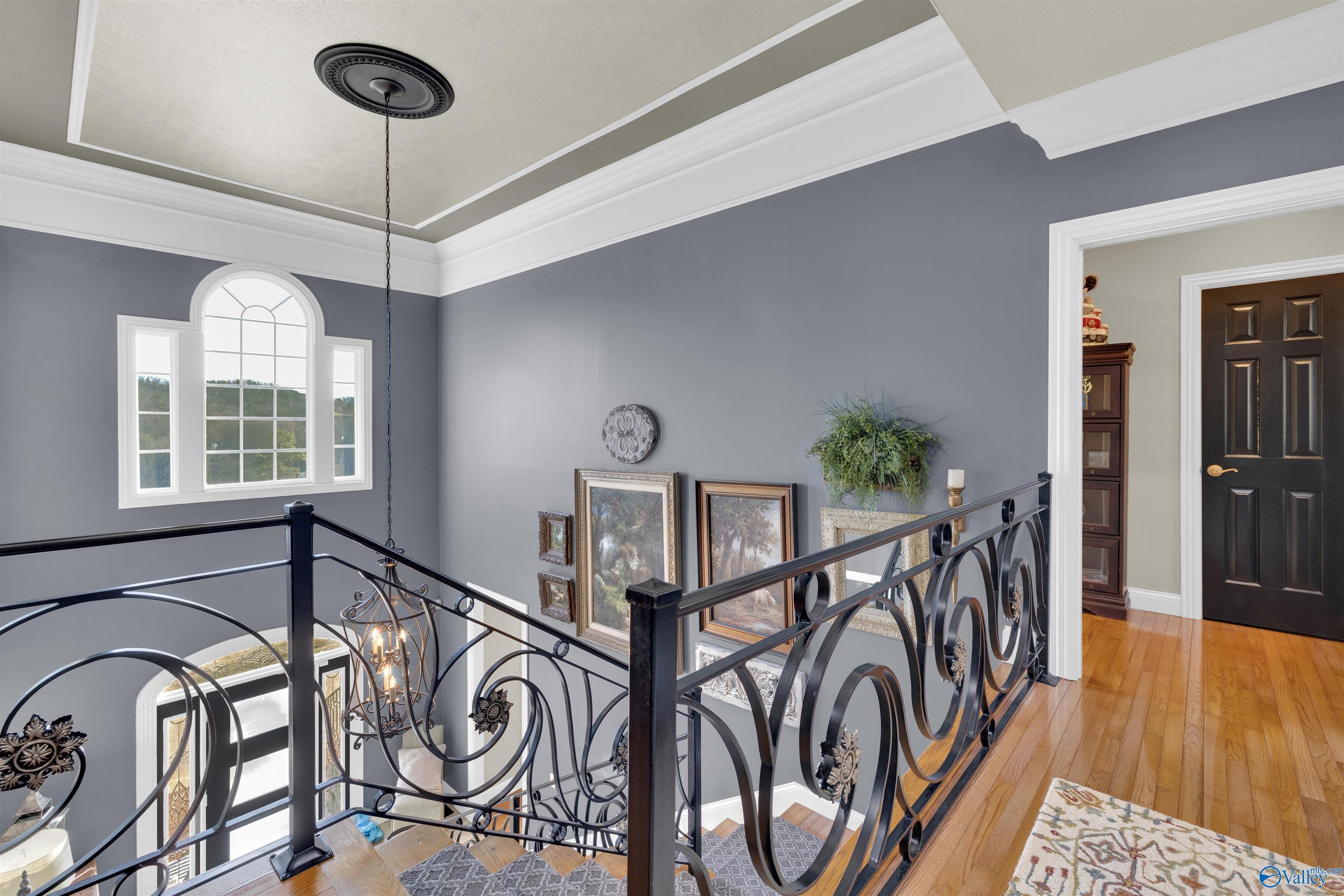 Upstairs landing with ornate black railings, an arched window, framed art, and hardwood floors.