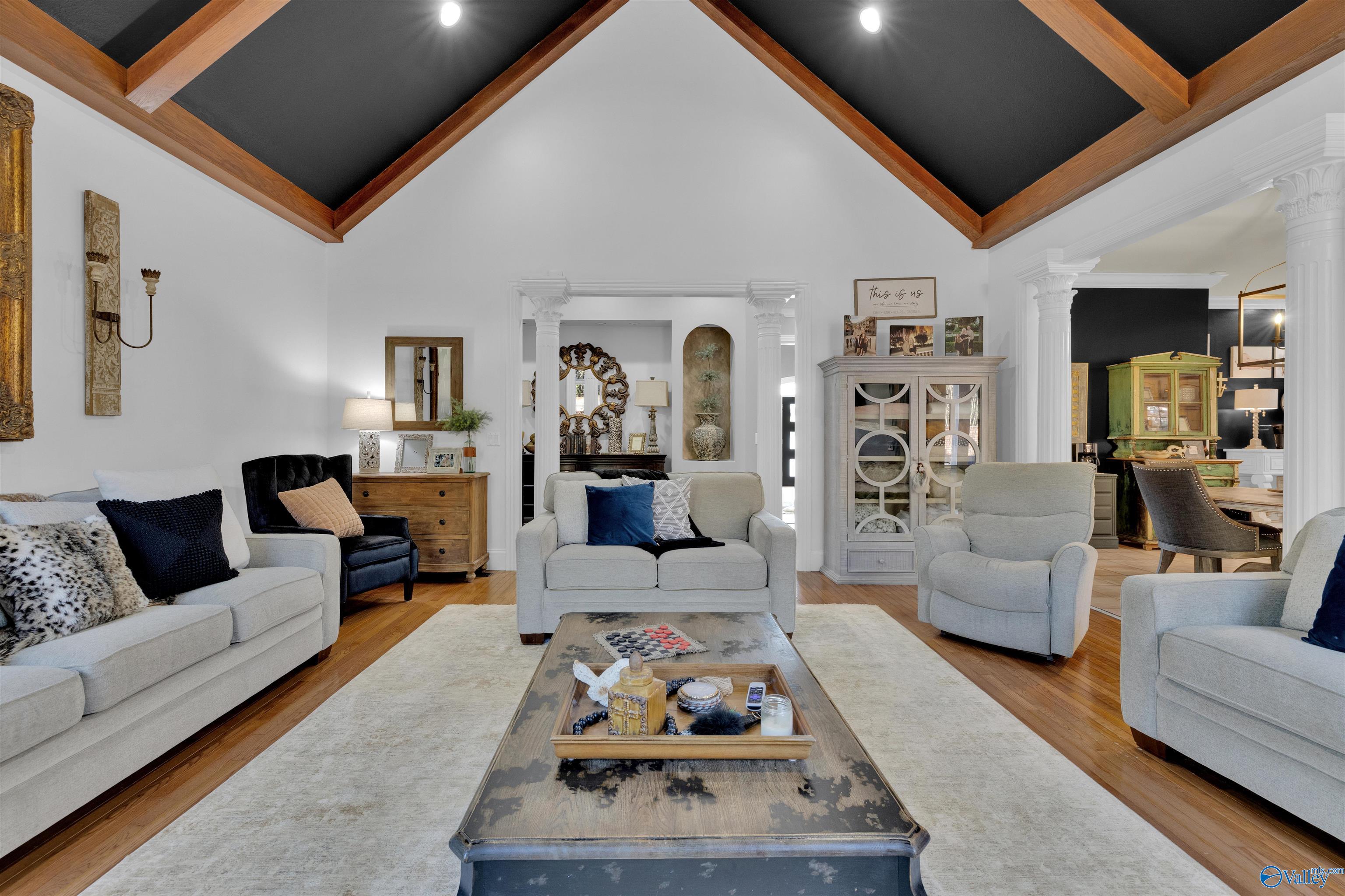 Living room with a dark vaulted ceiling, exposed wood beams, white columns, and light upholstered seating.
