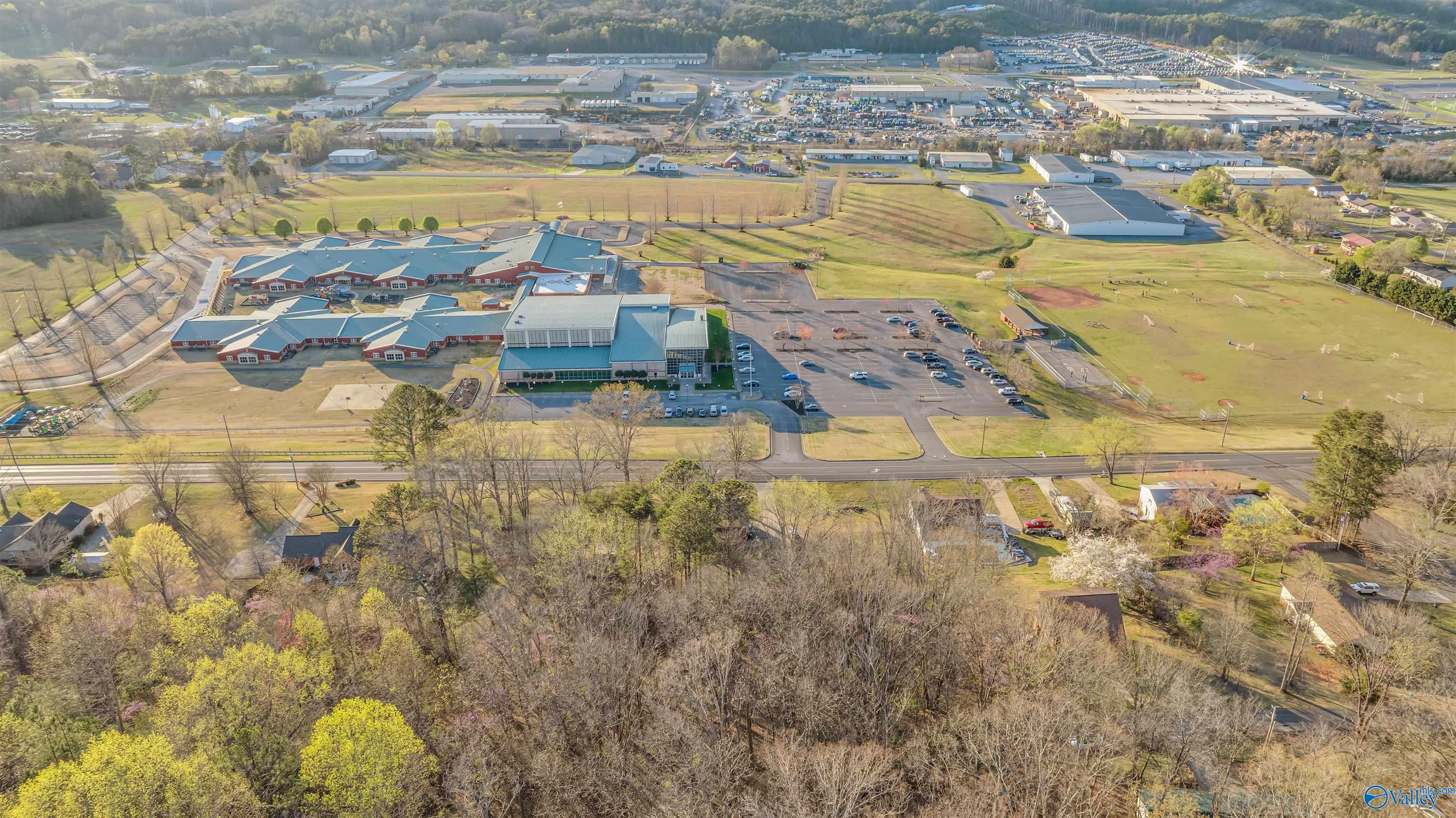 Aerial view of a school complex, sports fields, and surrounding buildings beside wooded neighborhoods.
