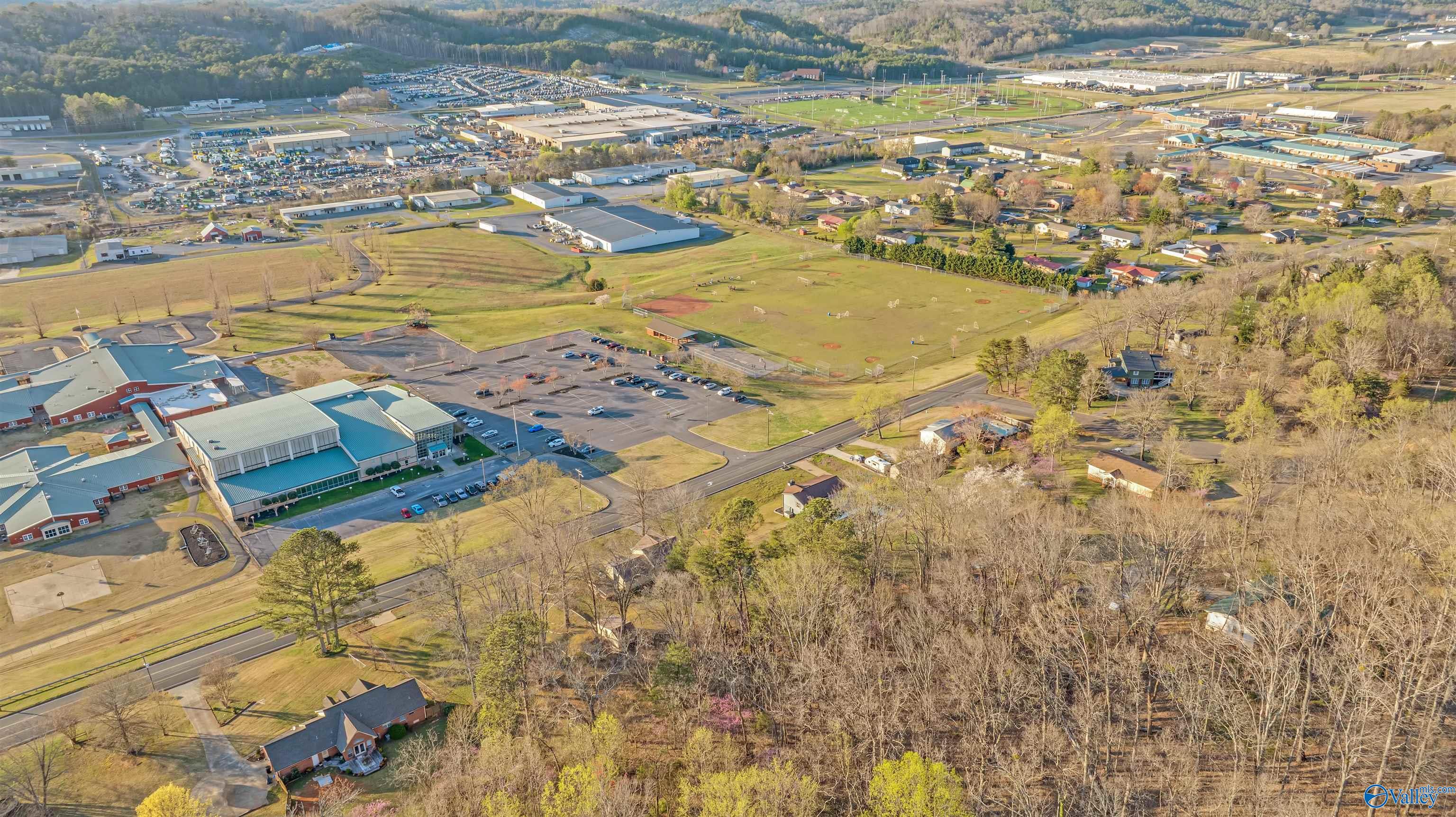 Aerial view of wooded land beside roads, homes, commercial buildings, and a large sports field.