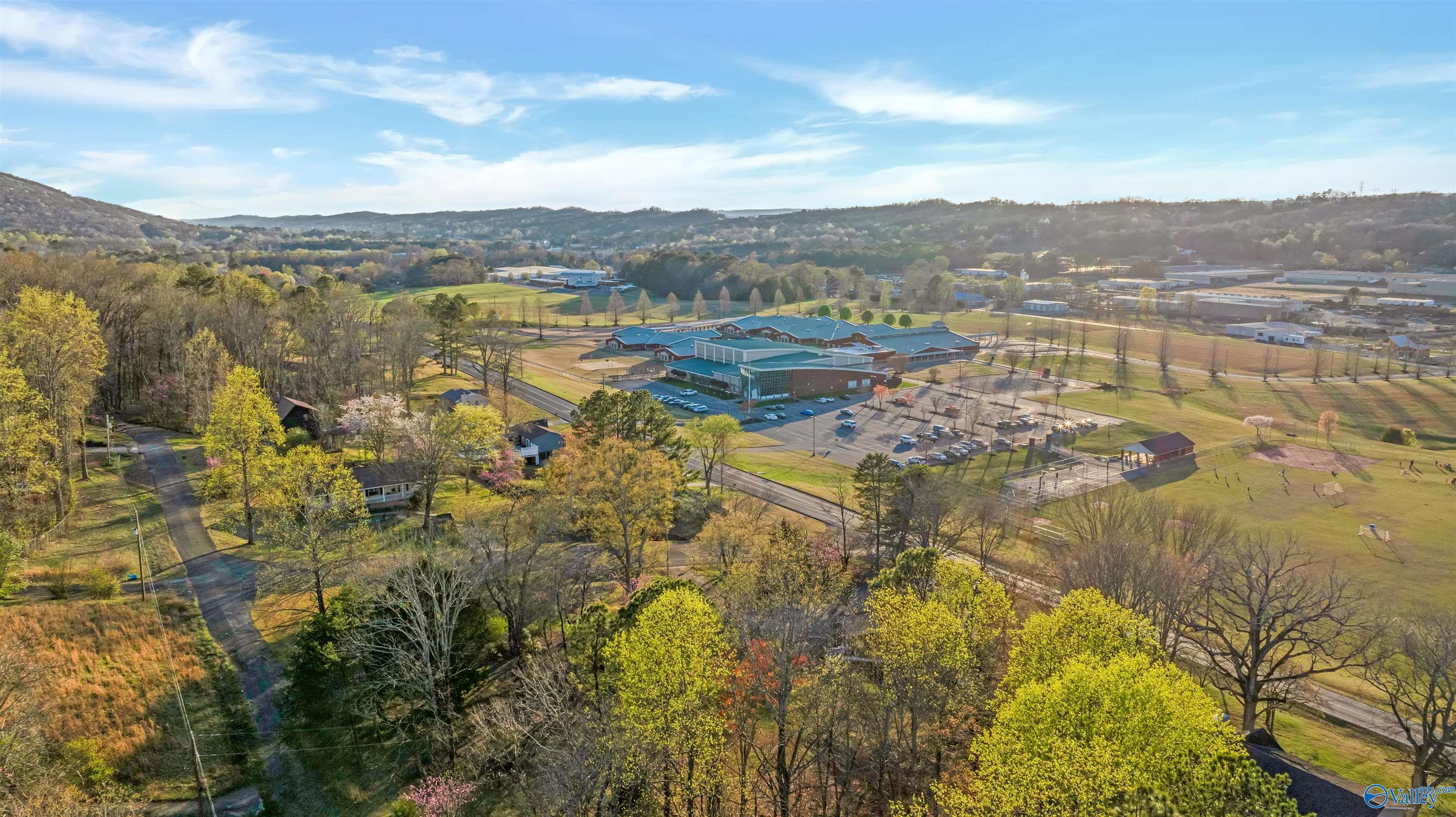 Aerial view of wooded surroundings, open fields, and a large complex beneath distant rolling hills.