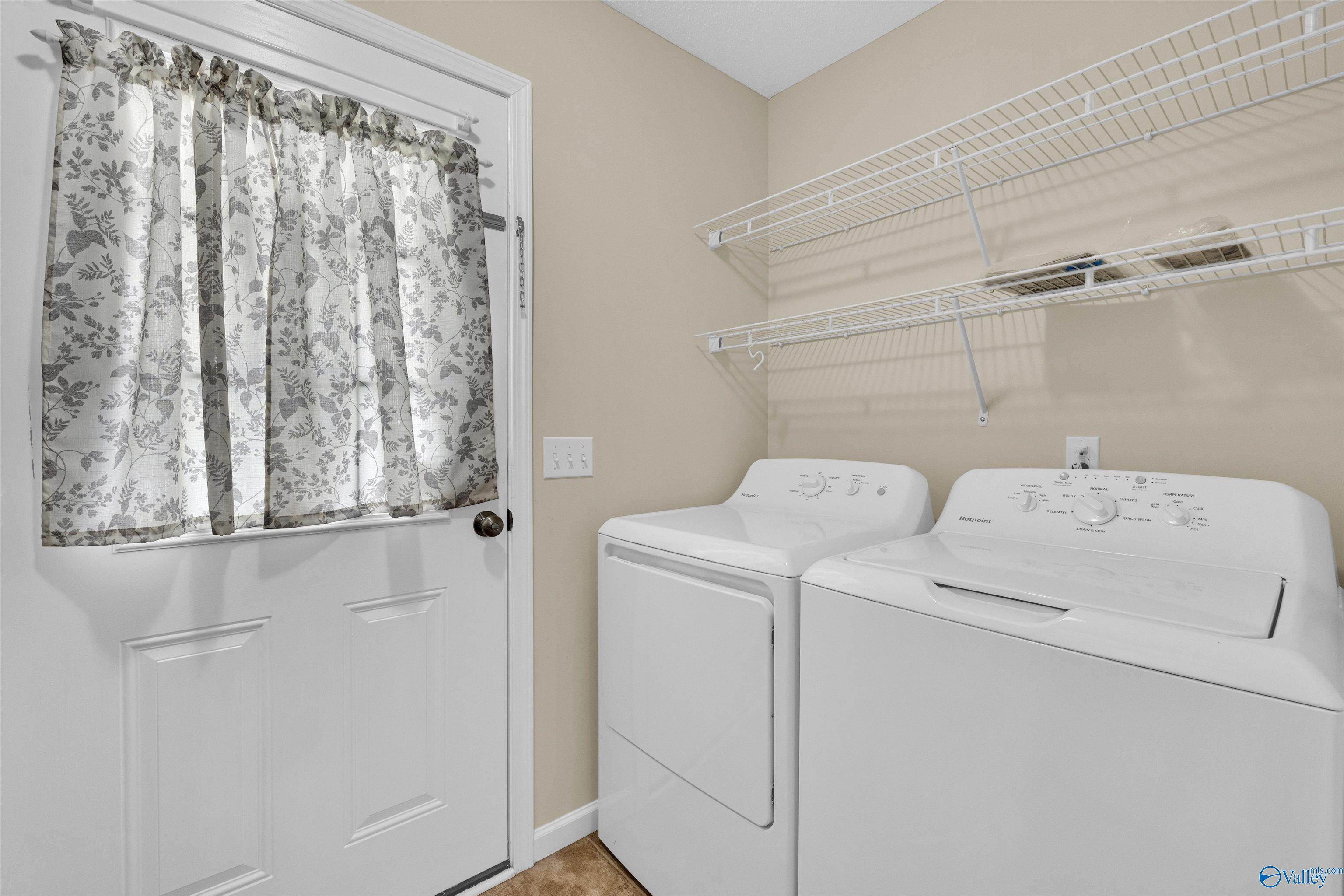 Laundry room with a white washer and dryer beneath wire shelves and a floral-curtained door.