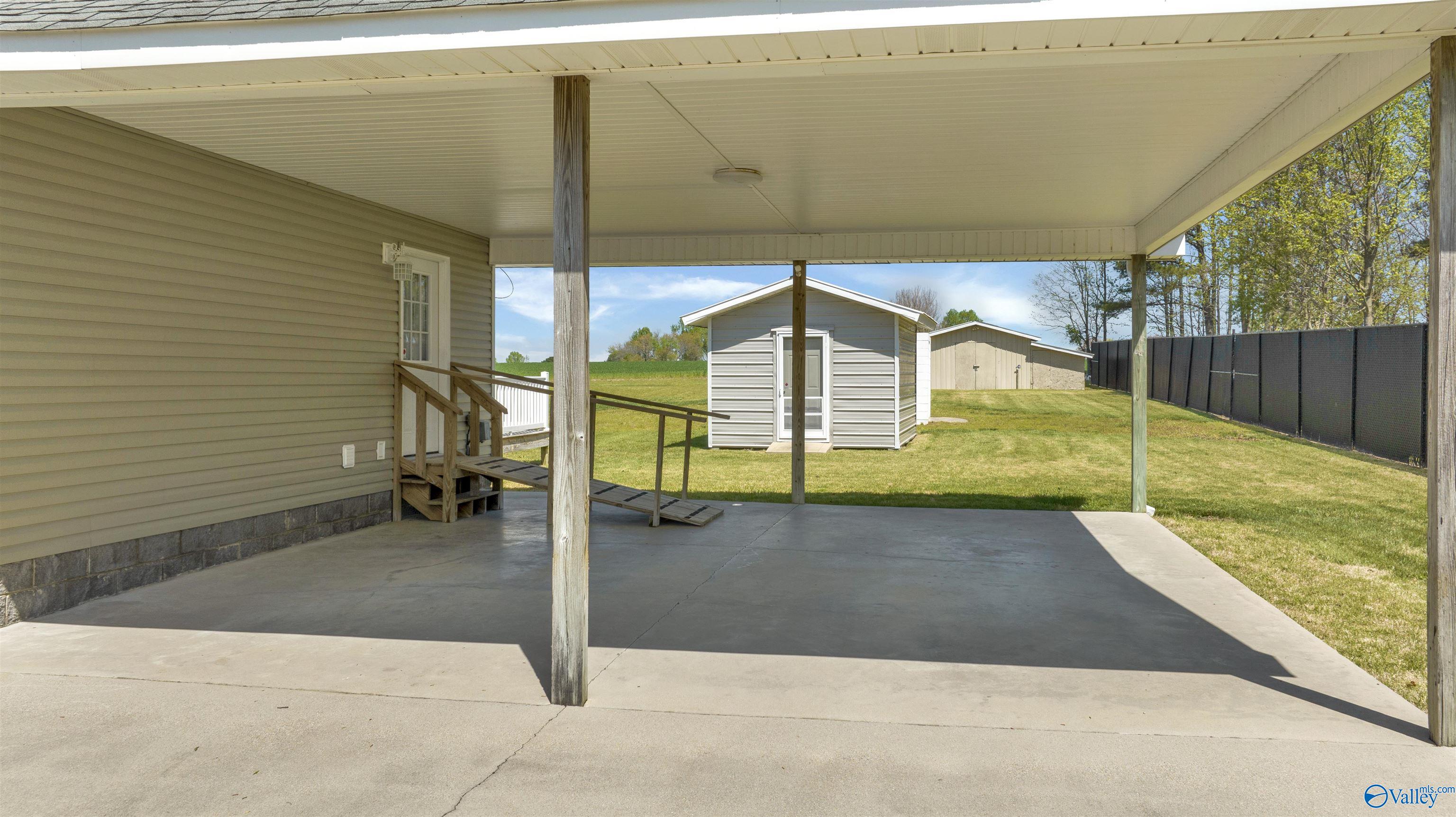 Covered concrete patio overlooks a fenced backyard with a shed and storage building.