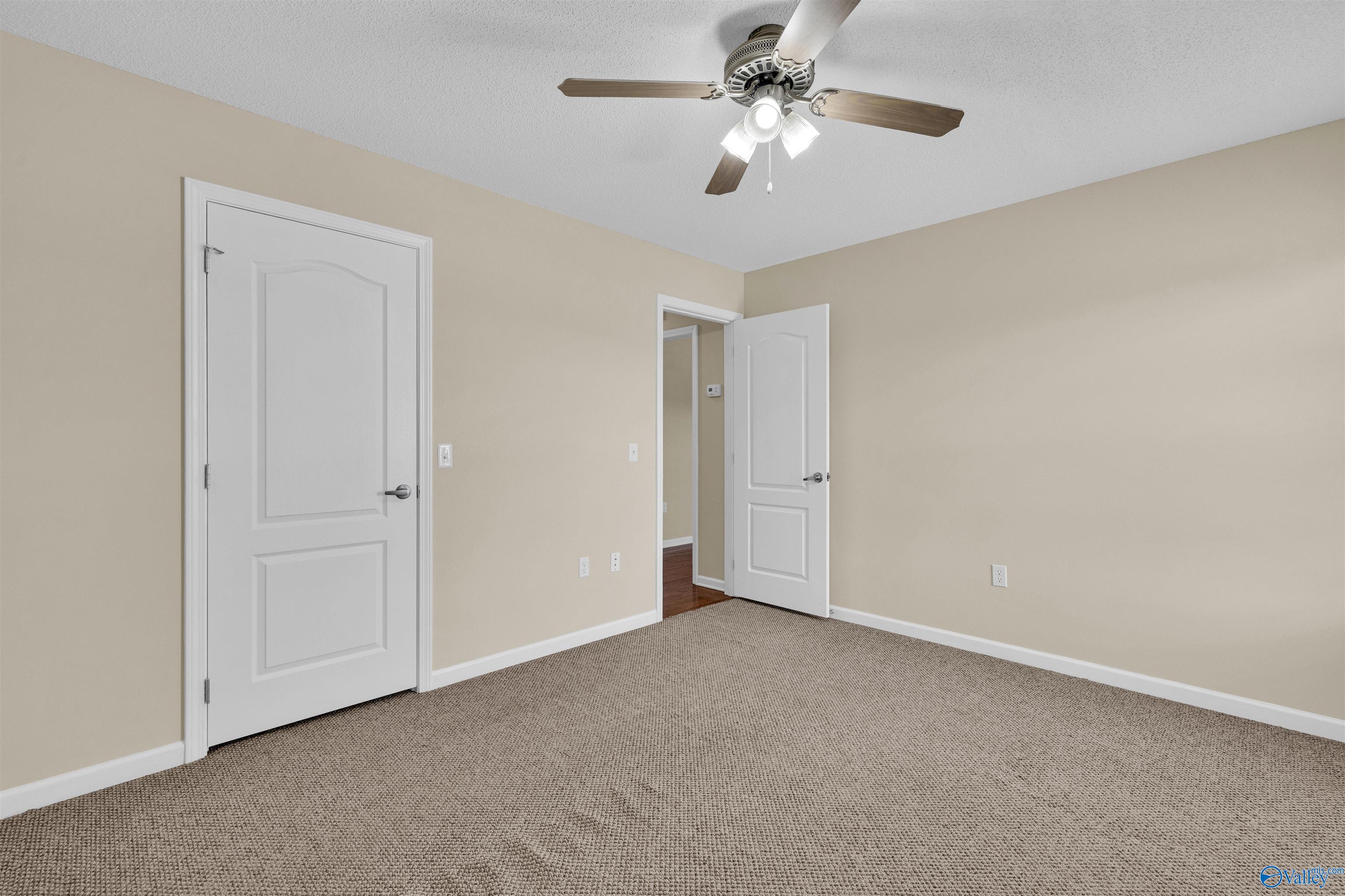 Empty bedroom with taupe carpet, beige walls, and a ceiling fan with light.