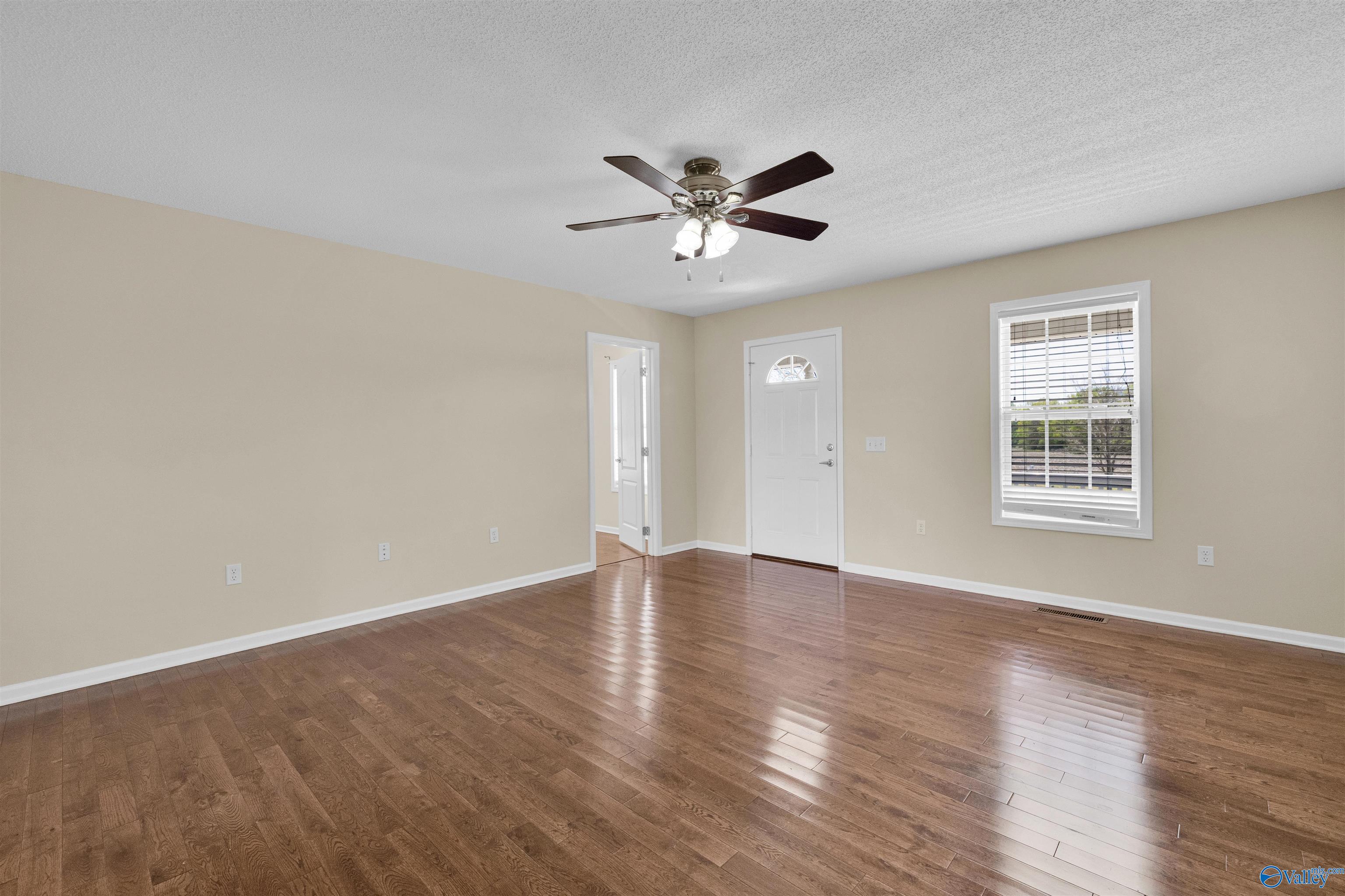 Empty room with glossy hardwood floors, a ceiling fan, front door, and window with blinds.