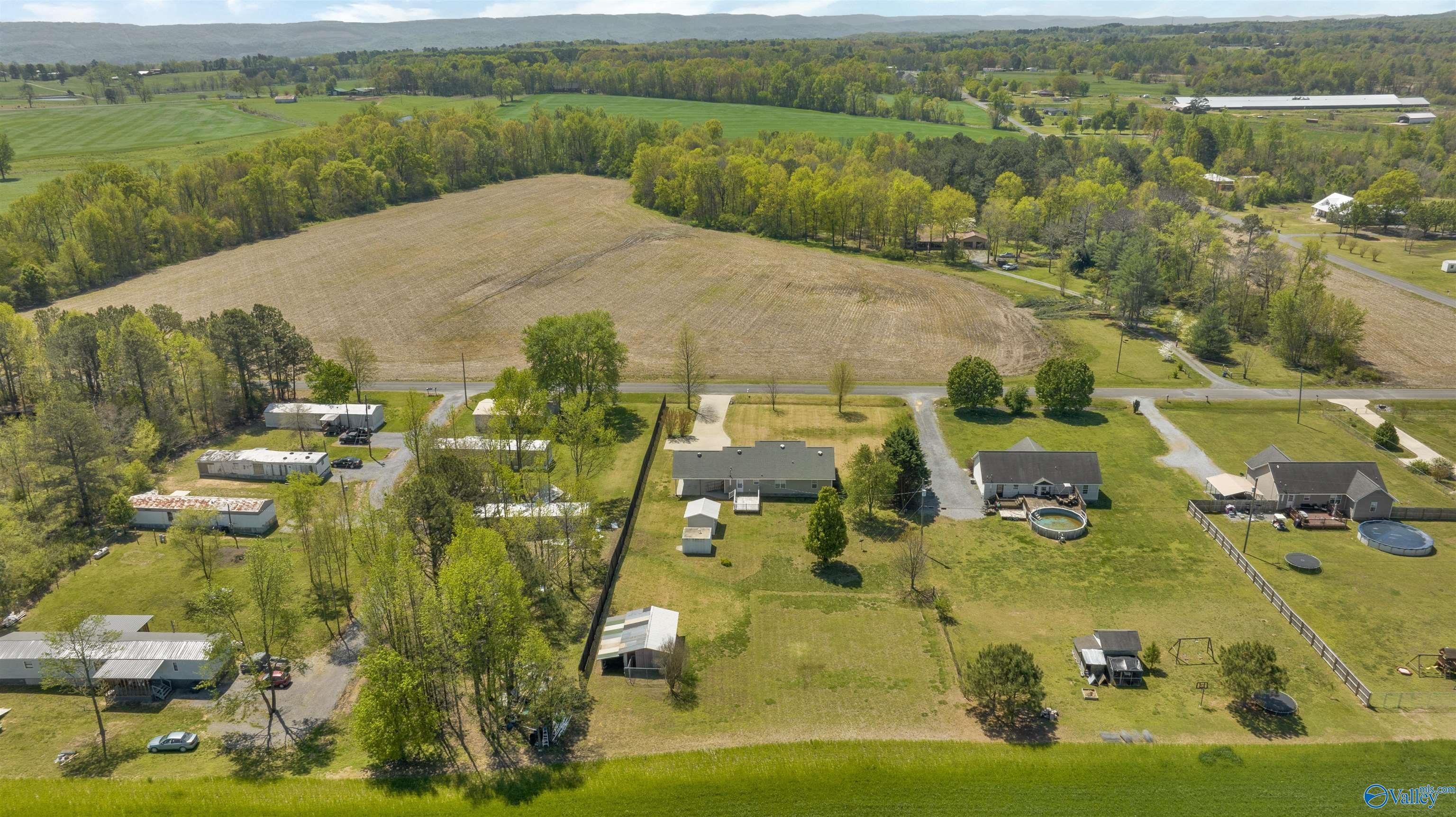 Aerial view of a rural home and outbuildings beside a large cultivated field and wooded hills.