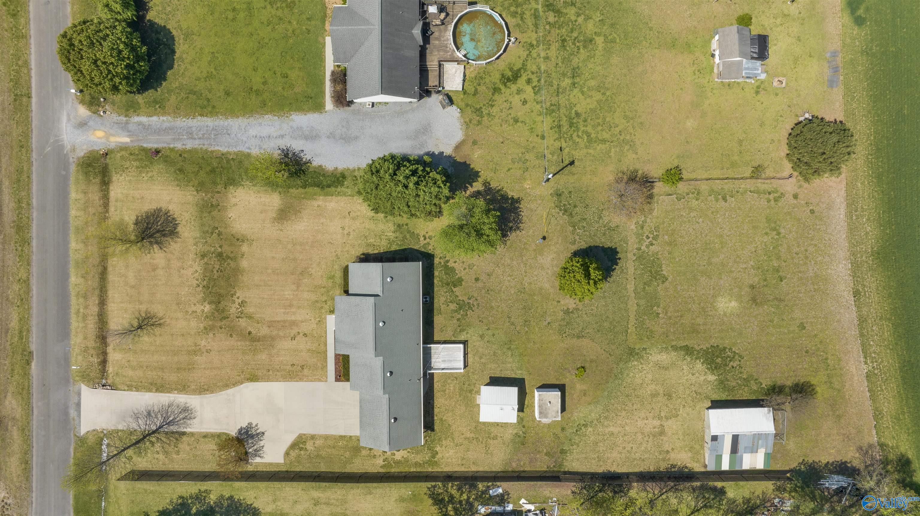 Aerial view of a house with a circular pool, long driveway, and several outbuildings across a large grassy lot.