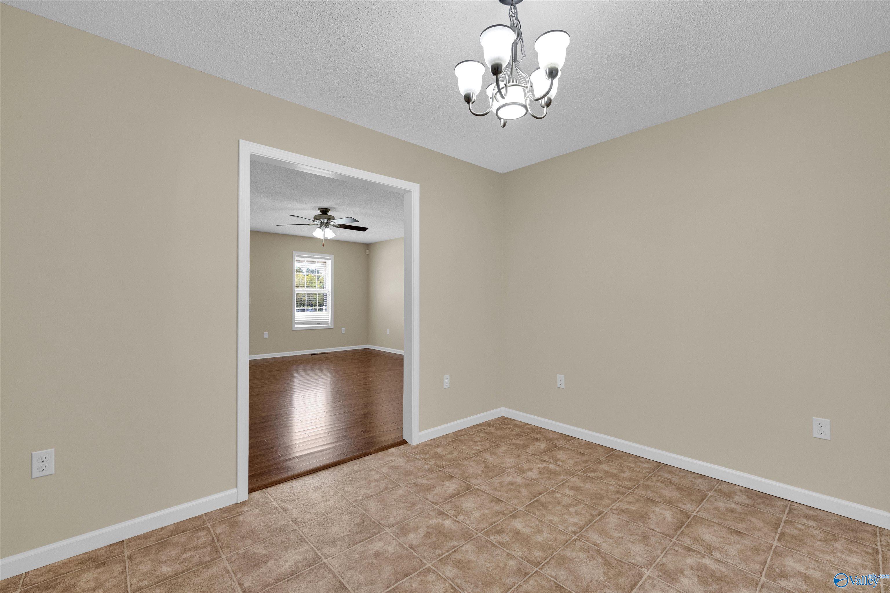 Empty beige room with brown tile flooring and a chandelier, opening to a hardwood-floored room.