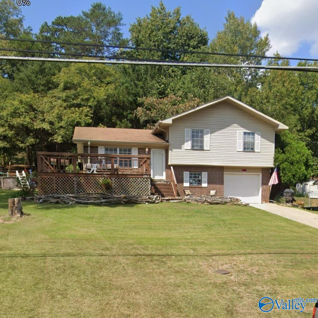Two-level beige and brick house with a raised deck, attached garage, and broad front lawn.