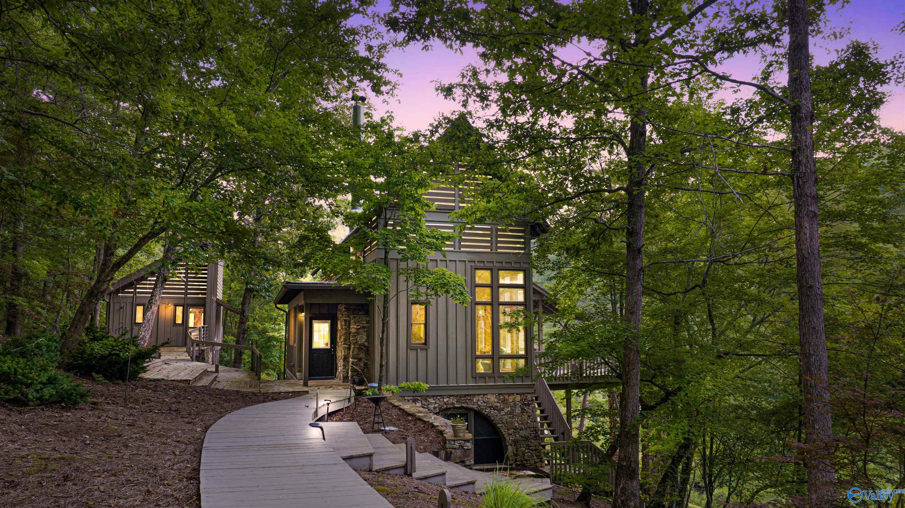 Modern multi-level home with glowing windows, stone foundation, and a curved walkway among trees at dusk.