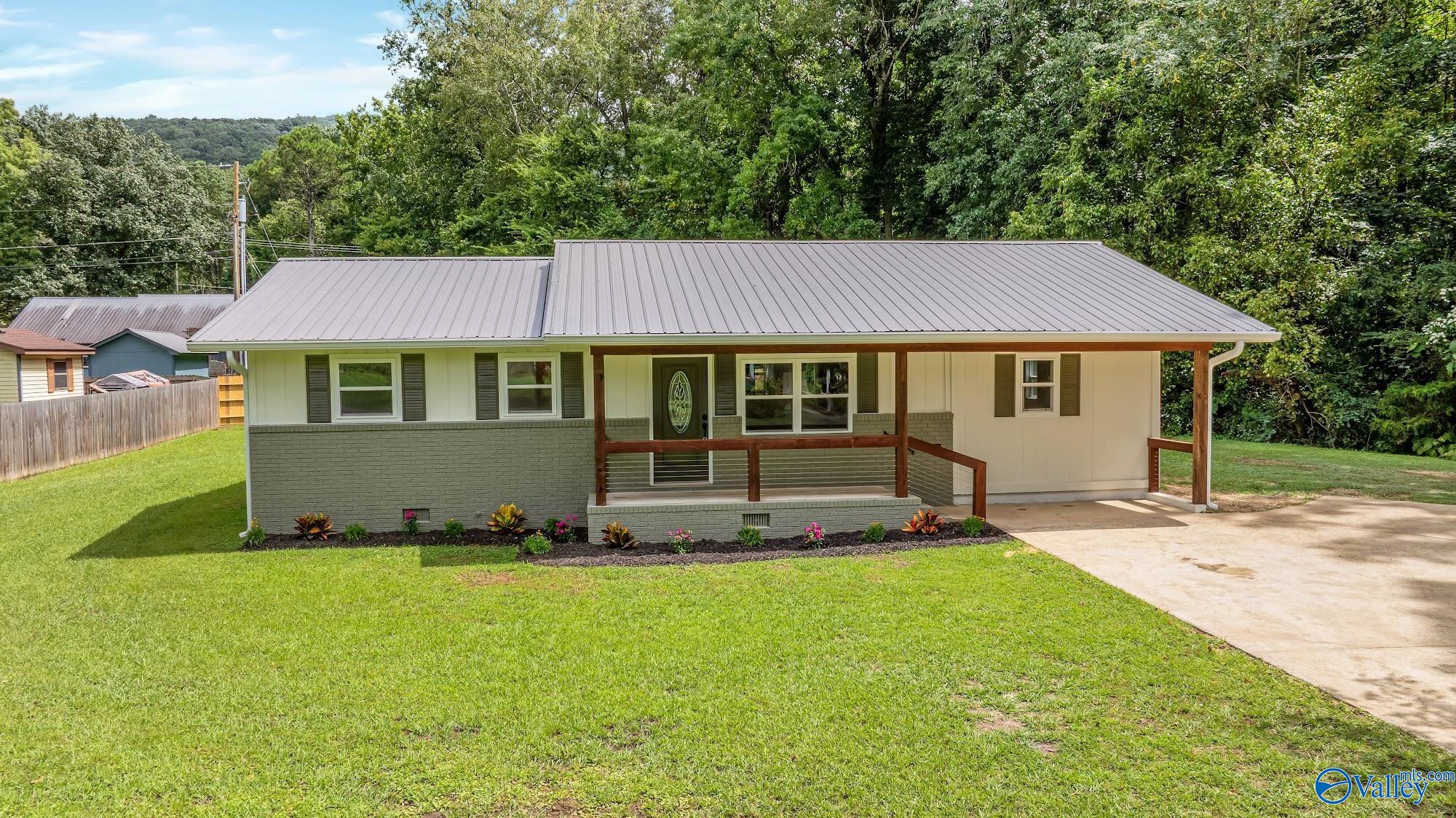 Single-story home with a metal roof, covered porch, landscaped lawn, and wooded backdrop.