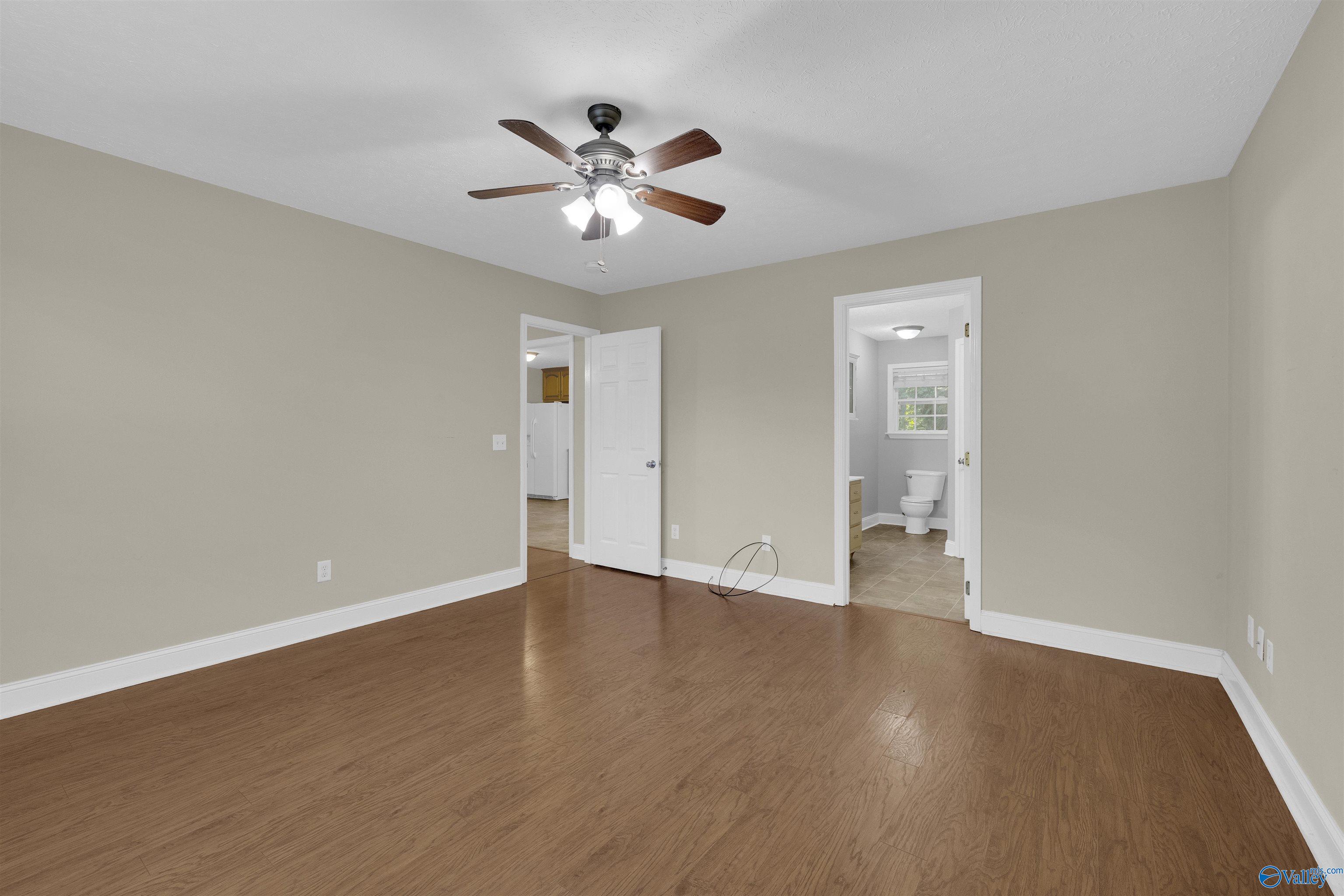 Vacant bedroom with wood-look flooring, a ceiling fan, and an open doorway to the bathroom.