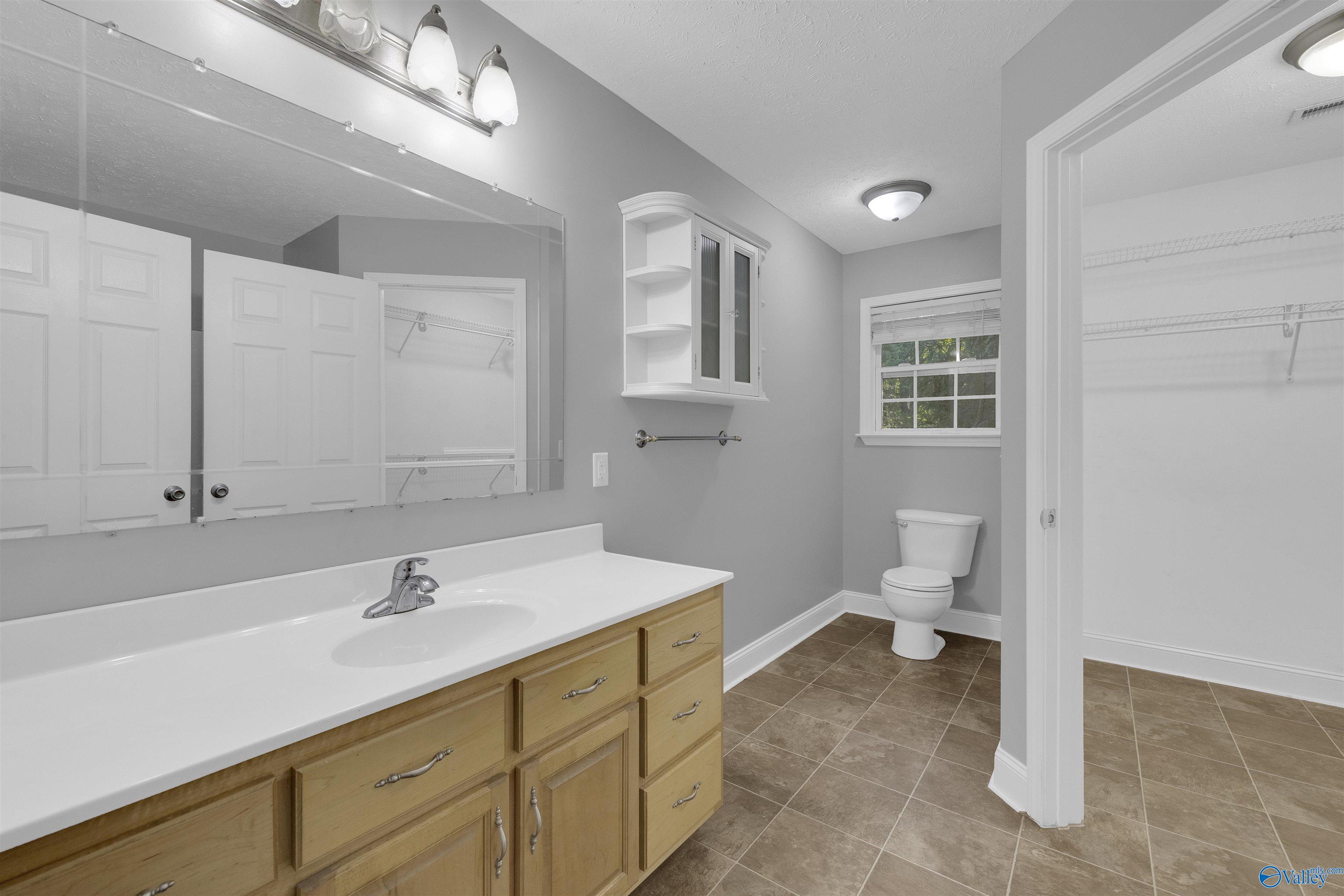 Bathroom with a long wood vanity, large mirror, toilet beneath a window, and an open closet doorway.