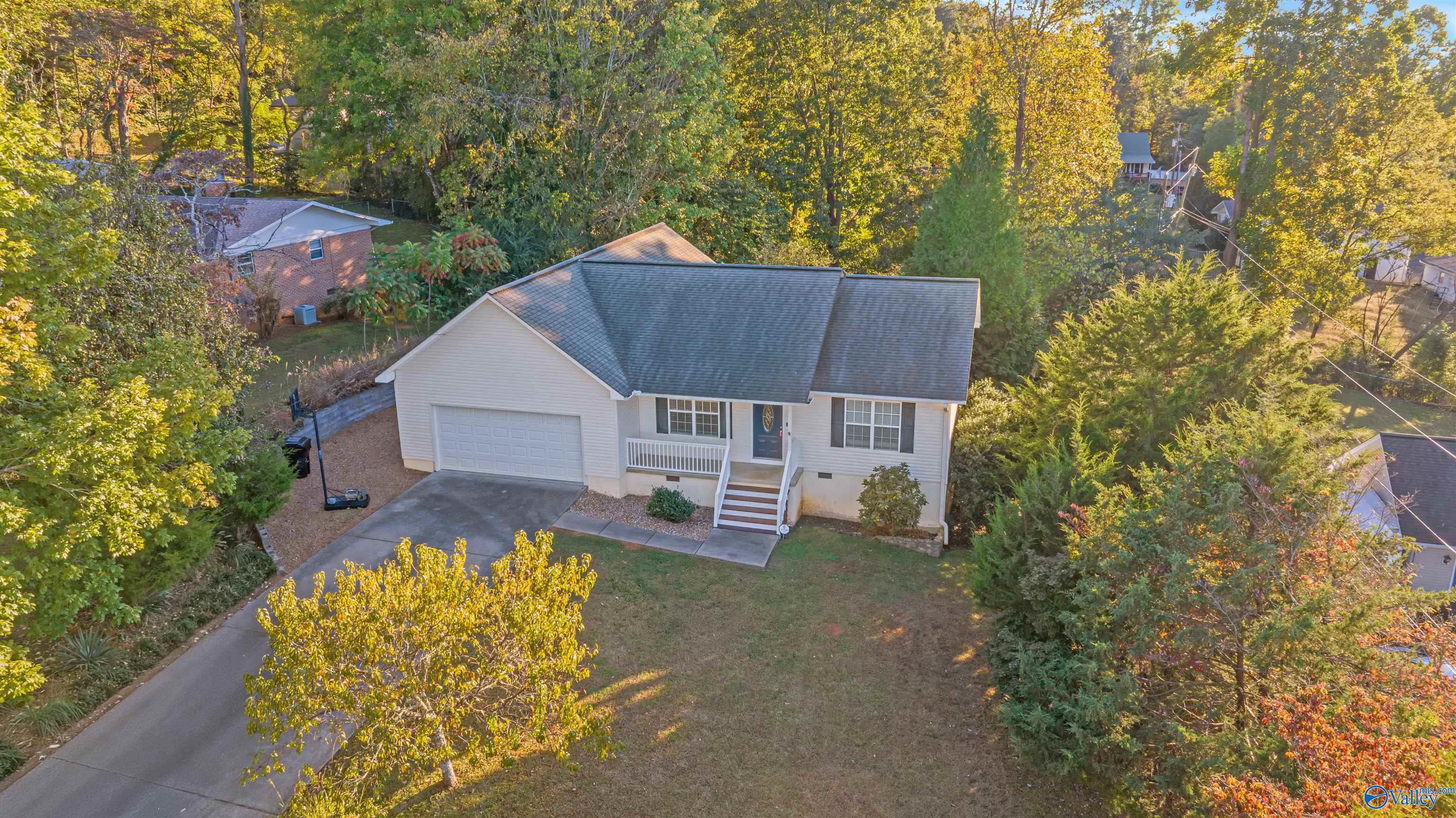 Aerial view of a light-colored home with an attached garage, driveway, and wooded autumn surroundings.