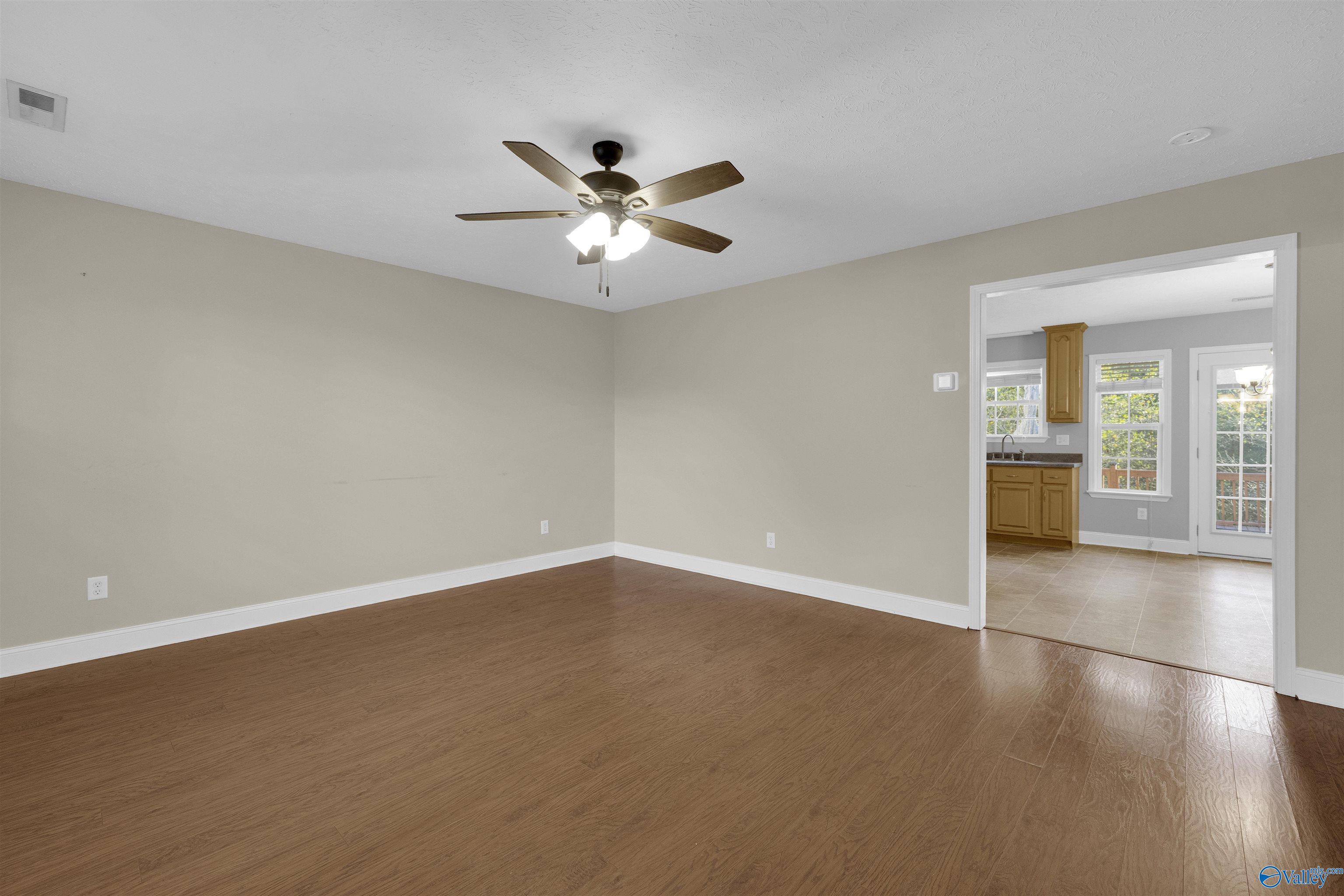 Empty room with wood-look floors and ceiling fan, opening to a tiled kitchen and breakfast area.