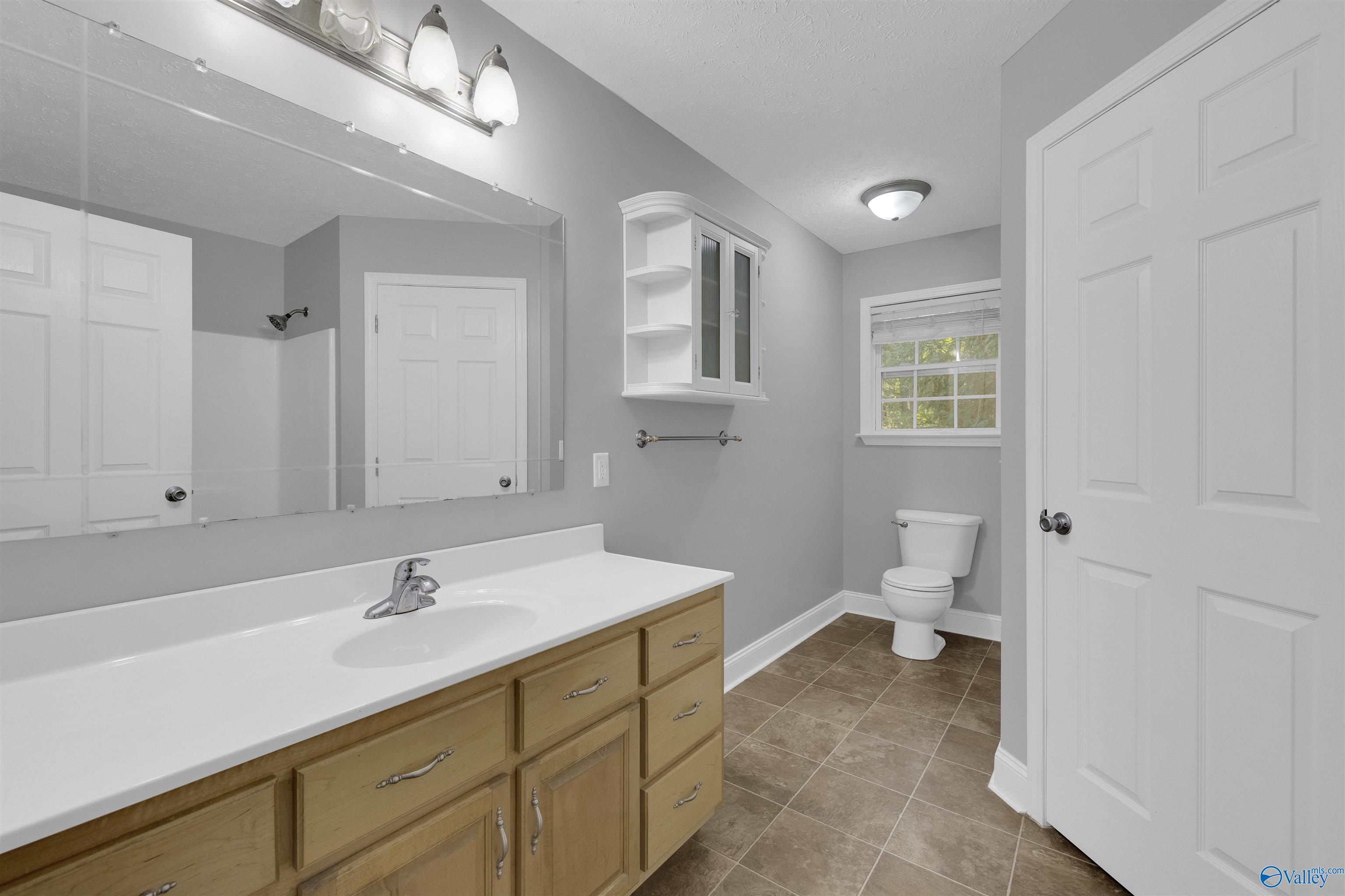Bathroom with a long white vanity, light wood cabinets, large mirror, and toilet beside a window.