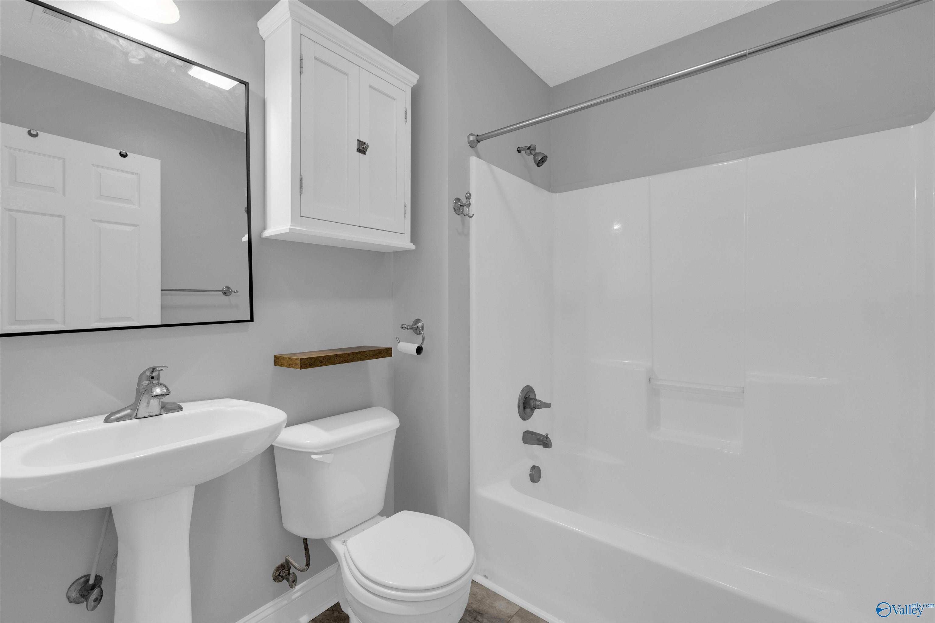 Bathroom with a white tub and shower, pedestal sink, toilet, and wall-mounted wooden shelf.