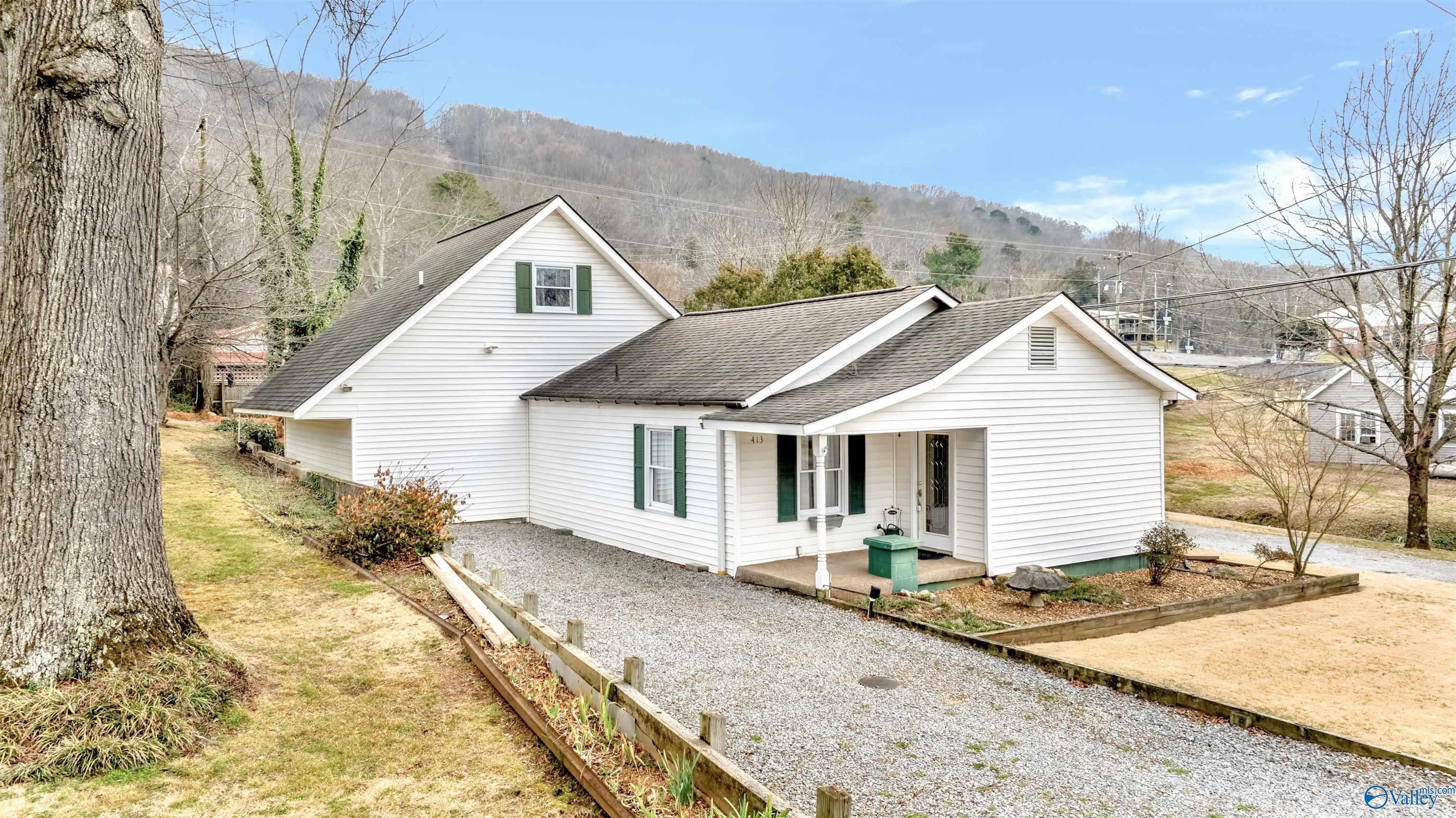 White gabled home with green shutters, gravel driveway, and wooded mountain backdrop.
