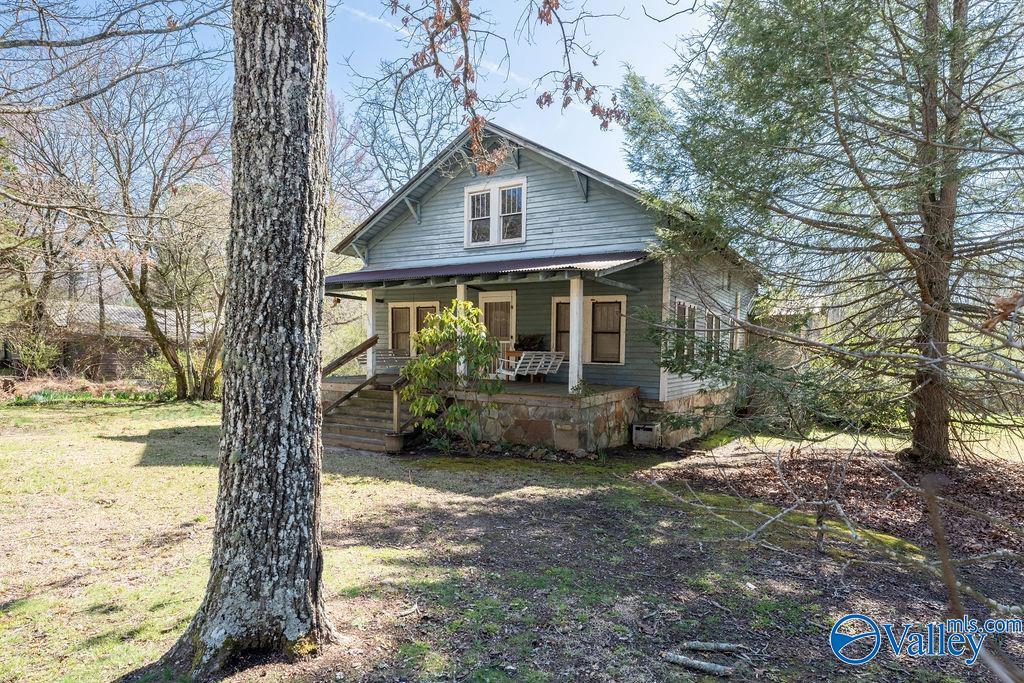Blue-sided house with a covered porch and stone foundation among mature trees.