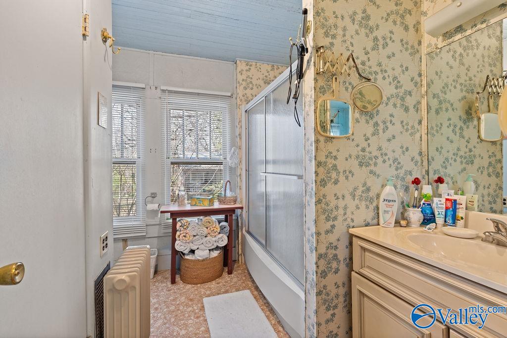 Bathroom with a glass shower, floral wallpaper, cream vanity, and towels arranged beneath a window.