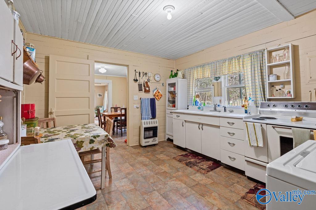 Kitchen with white cabinets, a double sink beneath curtained windows, and a dining table through the doorway.